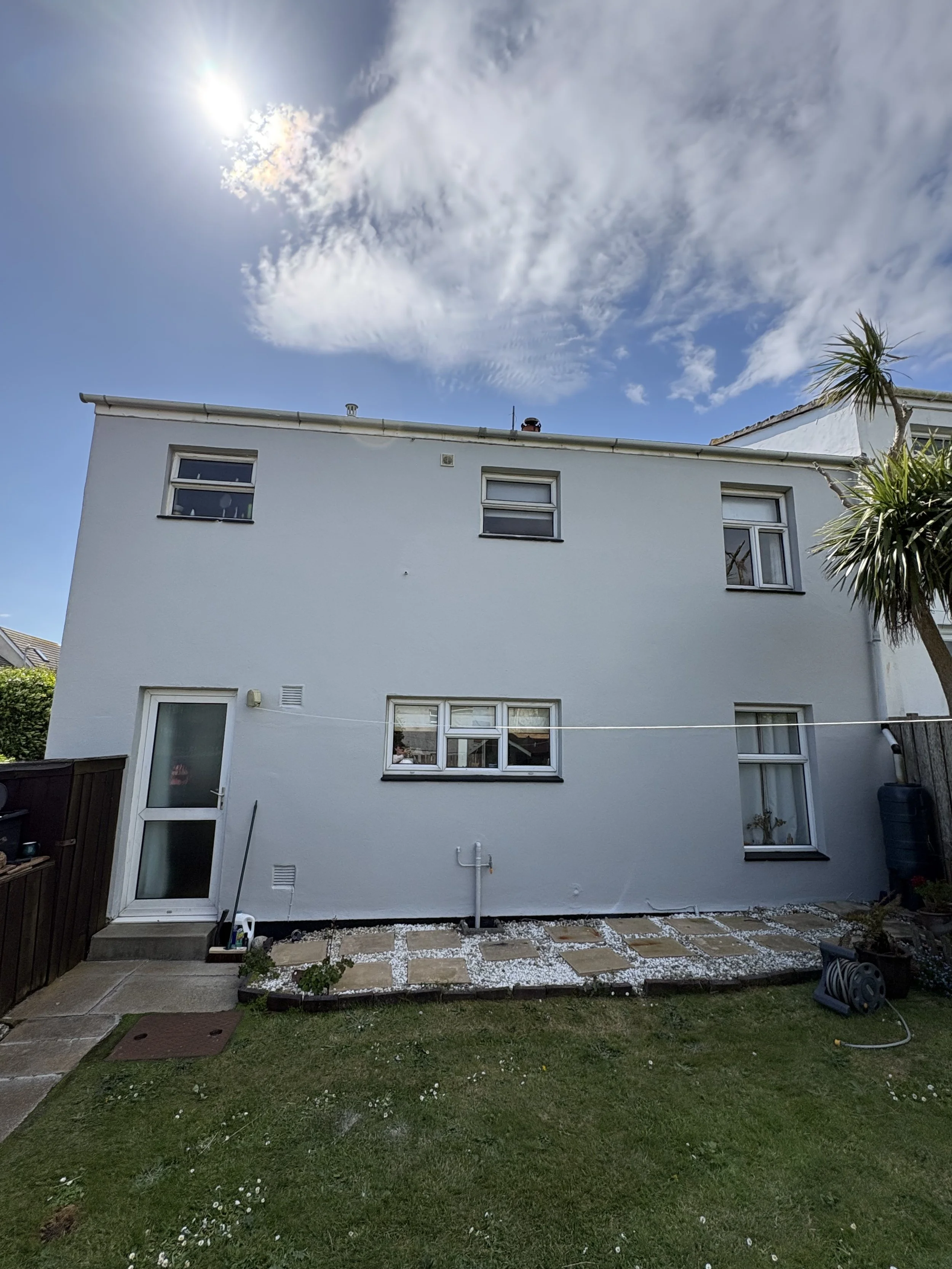 The back of a white house with three windows and a door, a small garden with a path of stones, and a green lawn. There are two trees, a palm tree on the right and a smaller tree on the left. The sky is mostly blue with some clouds and the sun shining brightly.