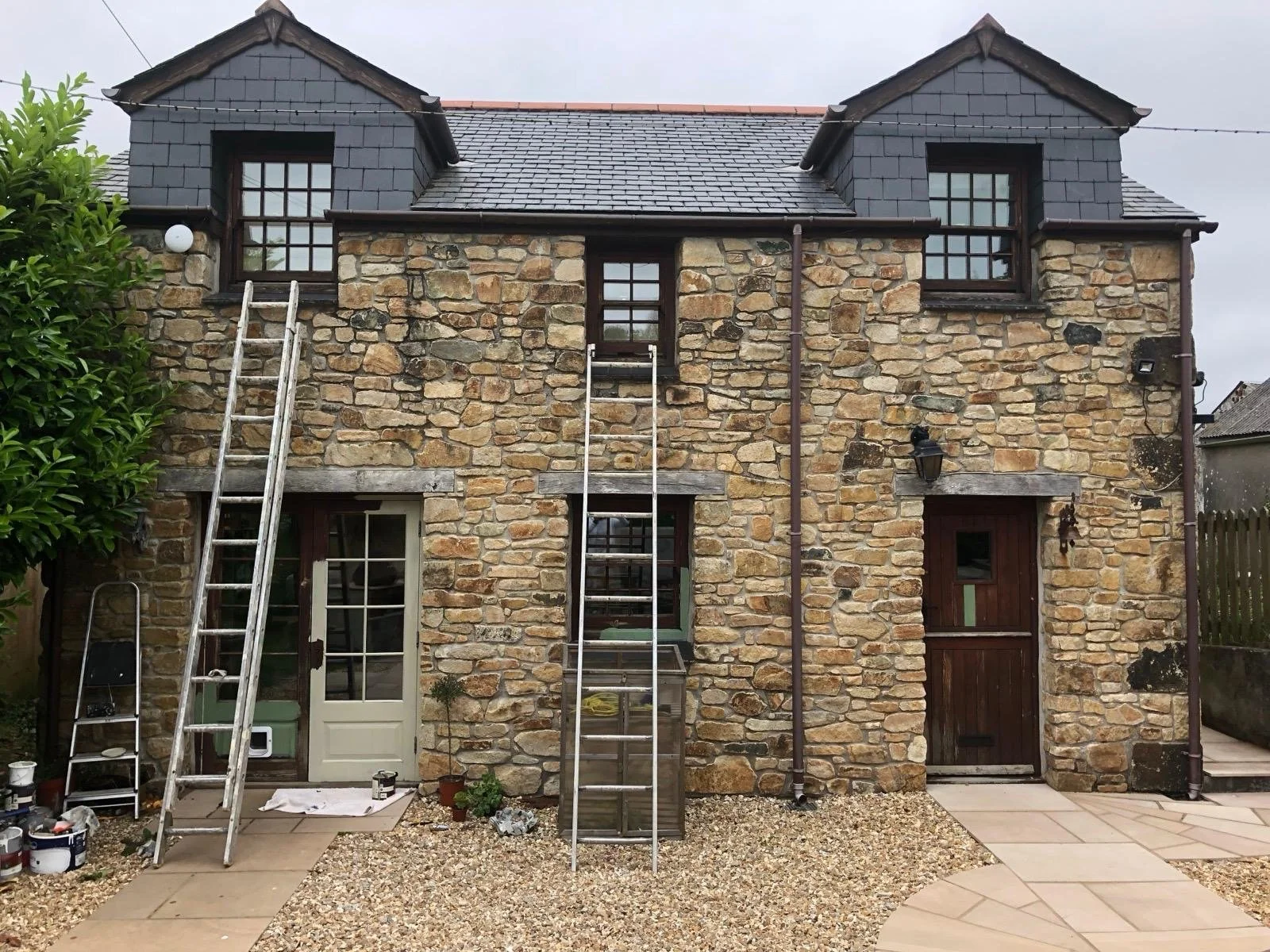 The back of a two-story stone house with black shingle roof, two dormer windows on the roof, and multiple windows on the second floor. Two ladders are leaning against the house, and nearby there are potted plants, paint cans, and construction equipment indicating ongoing renovation.