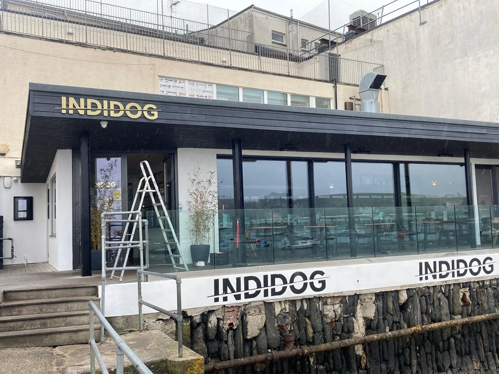 Exterior view of a modern building with large glass windows and black accents, with a sign reading 'INDIDOG' above the entrance and on a white barrier at the front, and a metal ladder leaning against the glass near potted plants.