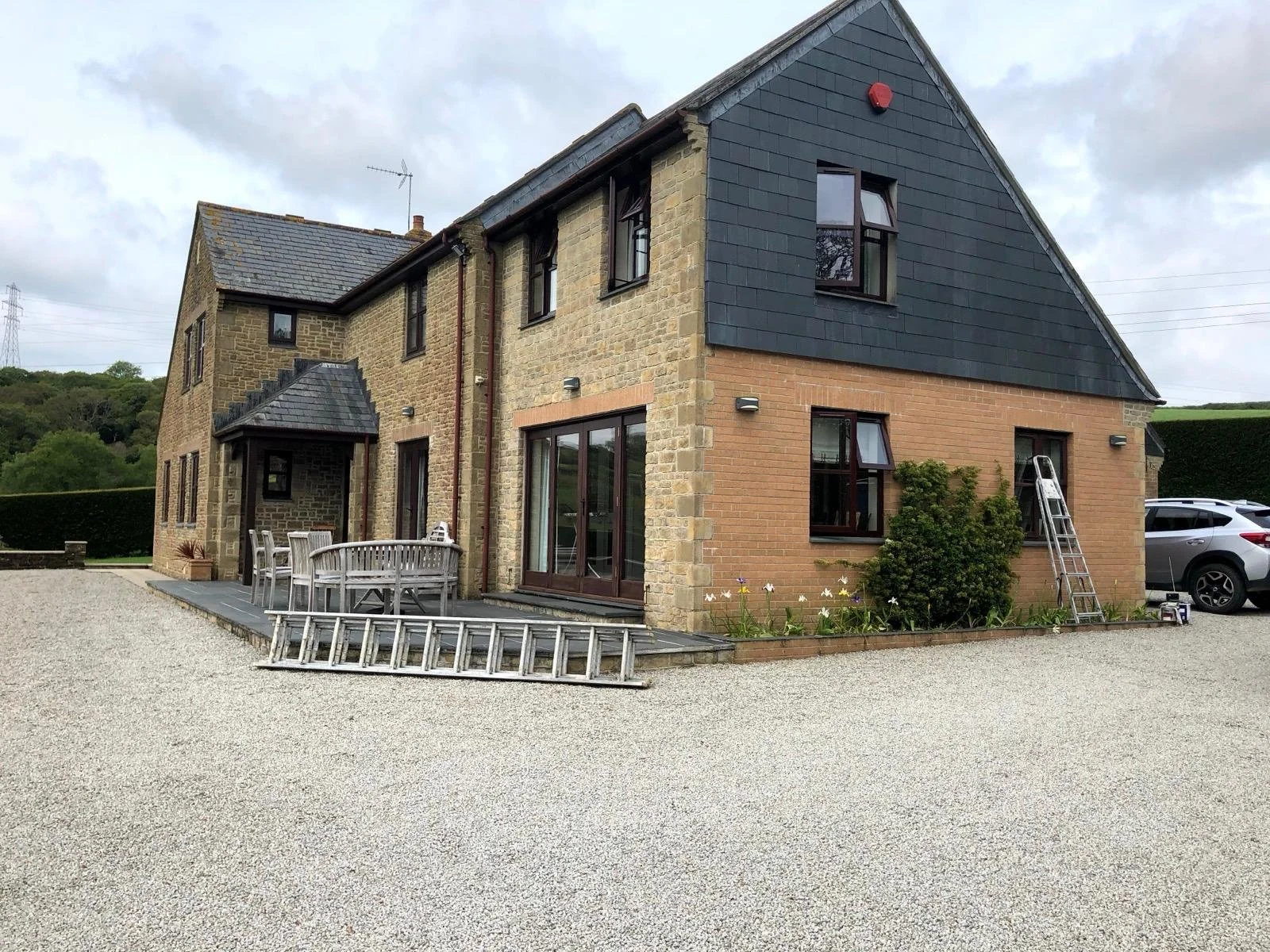 A multi-story house with stone and brick exterior, multiple windows, and a gravel driveway. There is a wooden outdoor dining table with chairs, a ladder leaning against the house, and a parking area with a gray SUV. Surrounding the house are green hills and cloudy sky.