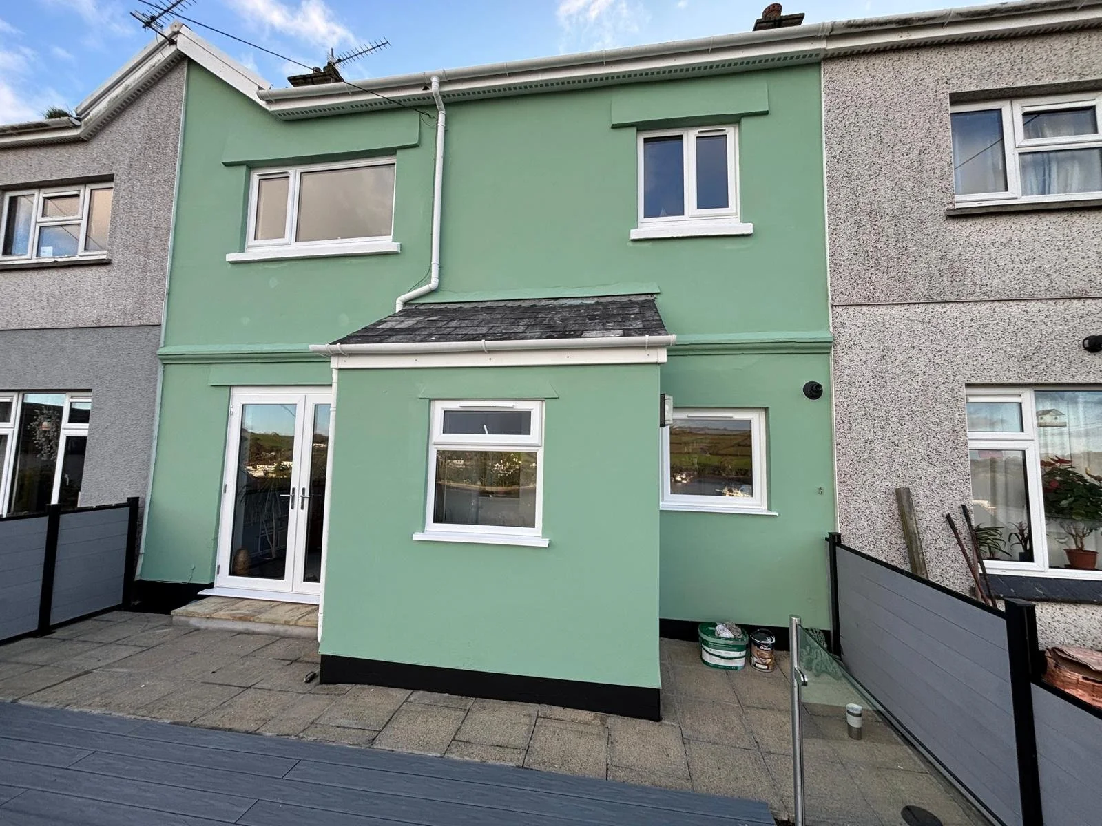 The back of a two-story house with a green exterior and white window frames, surrounded by a patio with paving stones and a black fence.