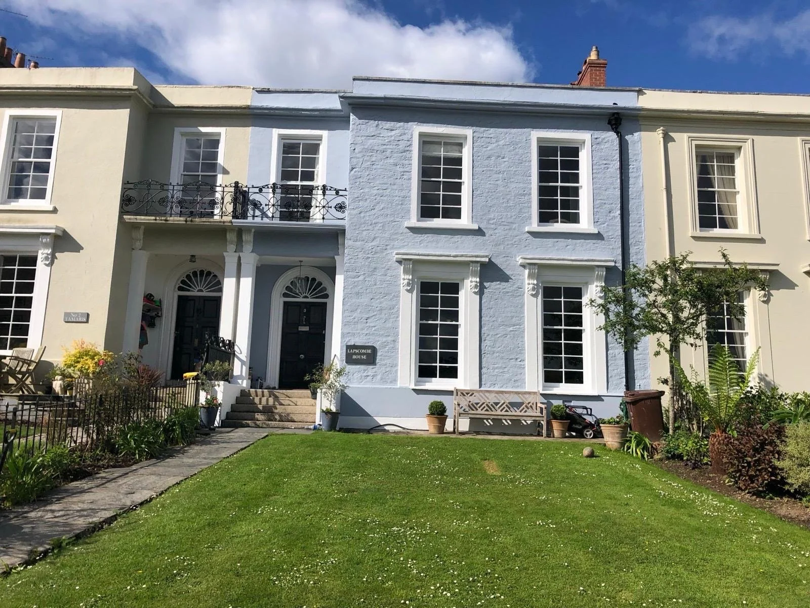 Row of attached houses with front gardens, black front doors, multiple windows, and a lawn in front. One house is painted light blue, while others are cream or beige. There are potted plants, a bench, and garden decorations in the front yard.