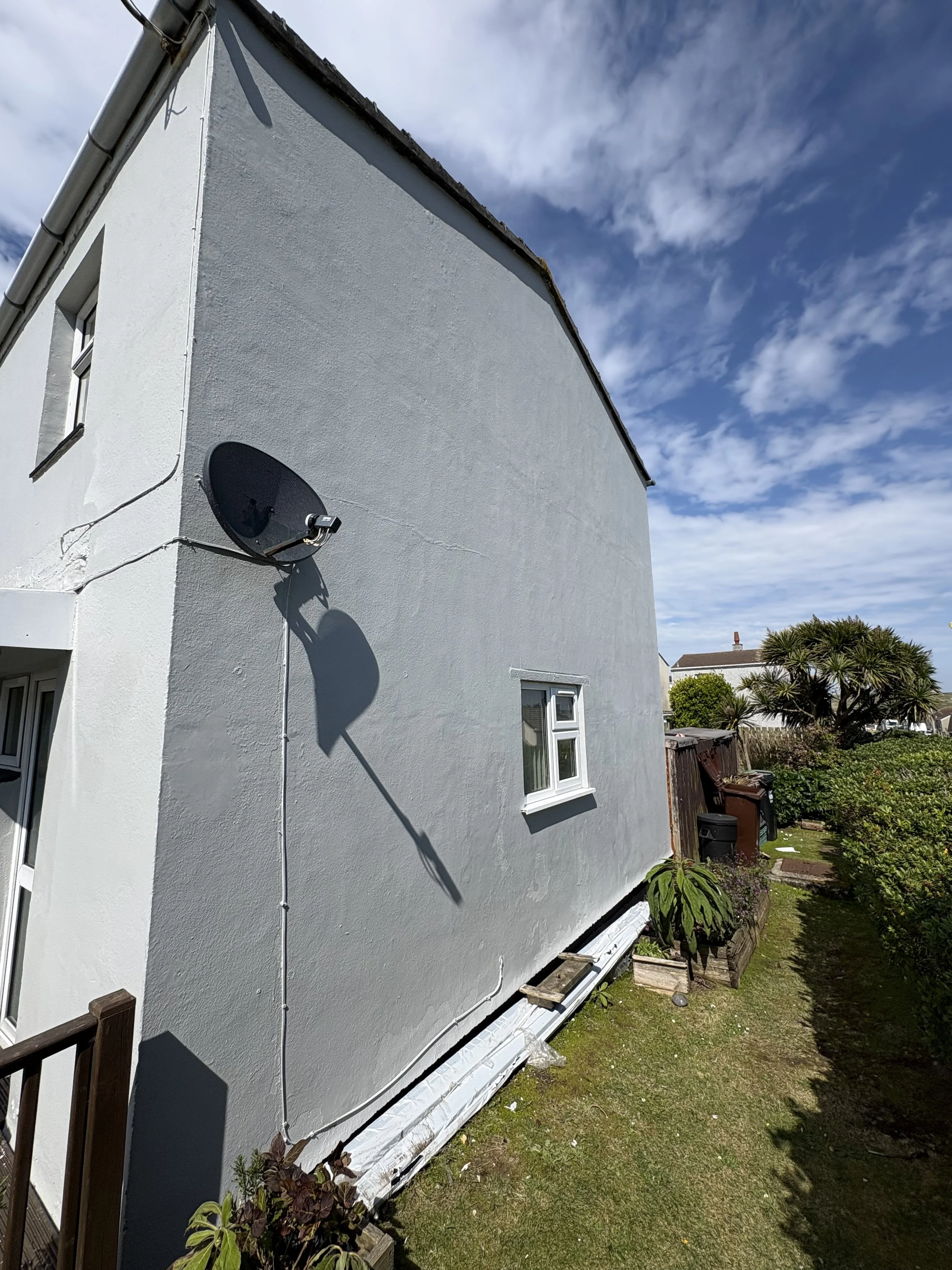 The side of a white house with a small window and a satellite dish, taken on a partly cloudy day. There is a garden with plants and a wooden fence.