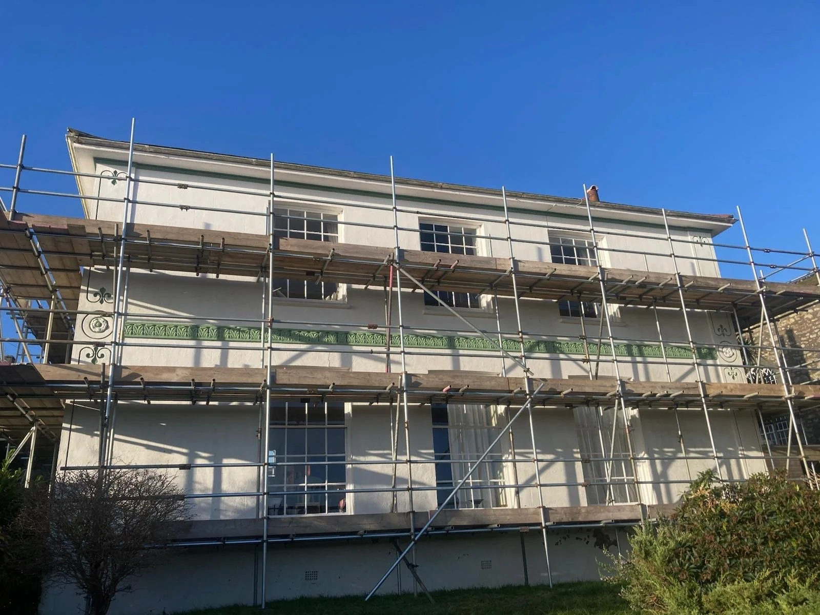 House with scaffolding around it, blue sky in the background, and some greenery at the bottom of the image.