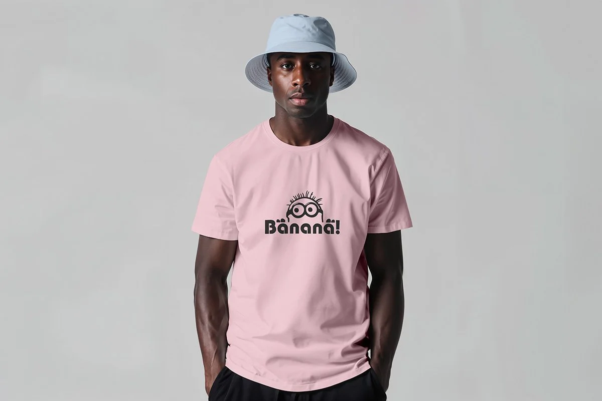 A young man standing against a neutral background, wearing a light pink T-shirt with a cartoon face and the word 'Bänaña!' printed on it, and a light blue bucket hat.