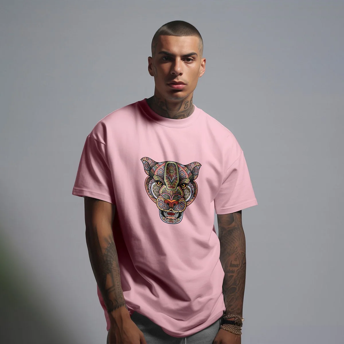 A young man with tattoos on his arms and neck, wearing a pink T-shirt with a colorful, decorative leopard face design on the front, standing against a plain gray background.