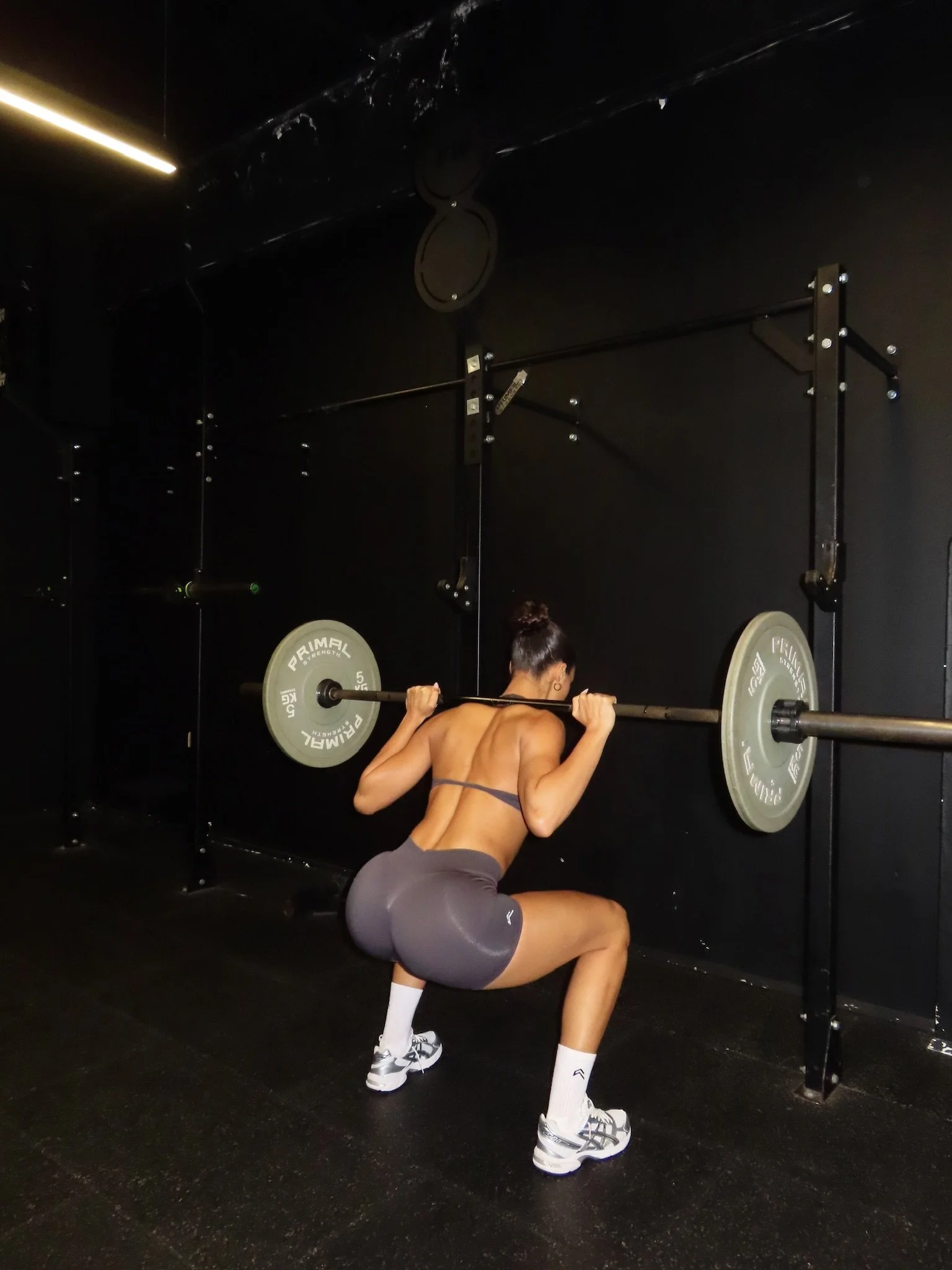A woman with dark hair in a bun performing a squat exercise with a barbell loaded with weight plates at a gym with black walls.