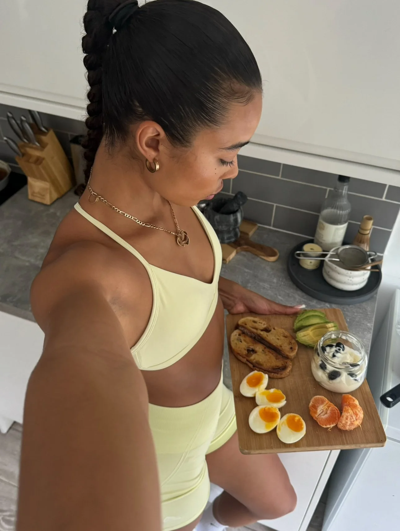 Woman in yellow sportswear taking a selfie in the kitchen with a breakfast tray containing boiled eggs, toast, avocado, blueberries in yogurt, and orange segments.