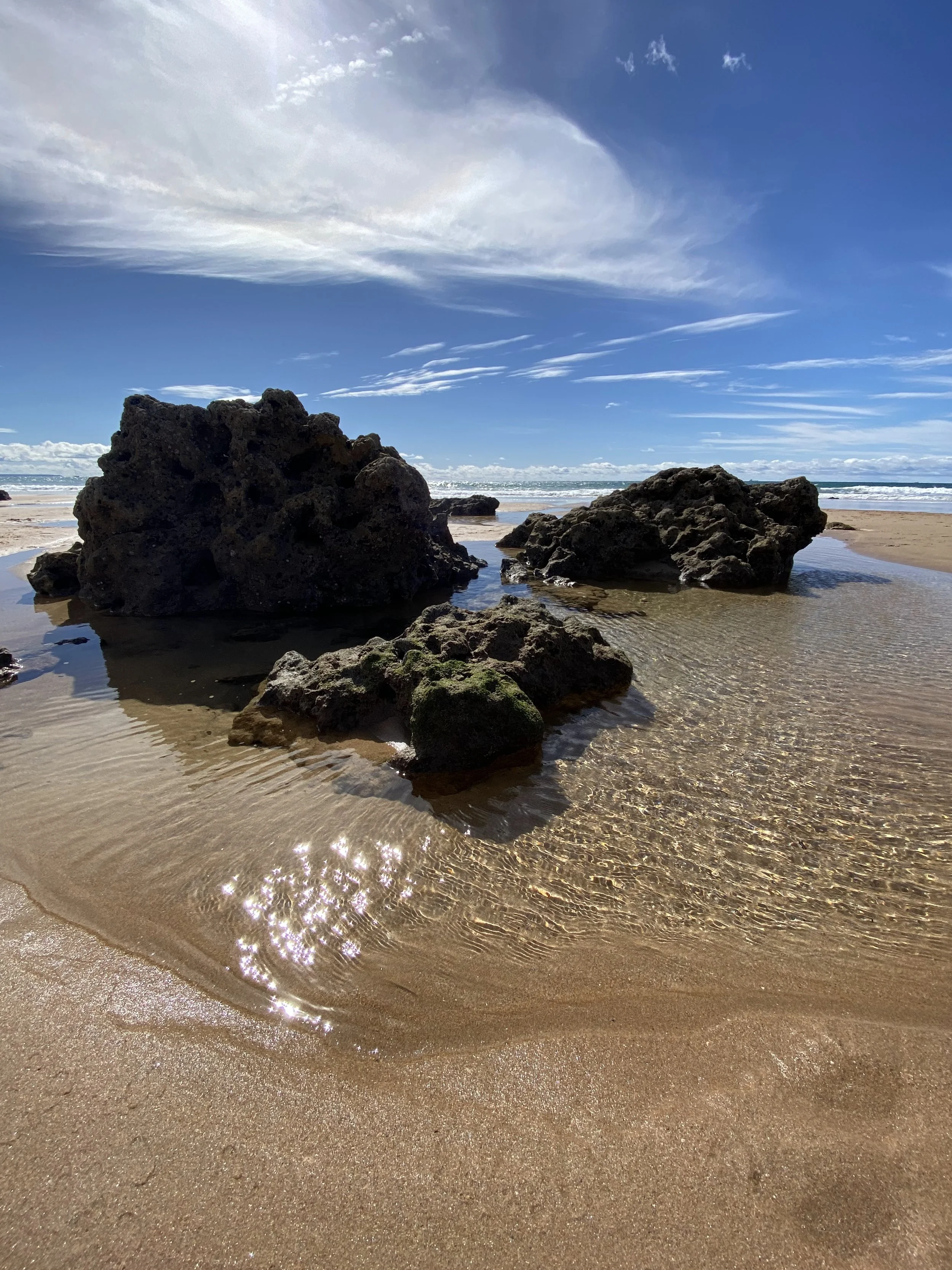Portuguese beach scene (Algarve) with large rocks on wet sand, gentle waves in the background, and a partly cloudy sky with white clouds and blue sky.