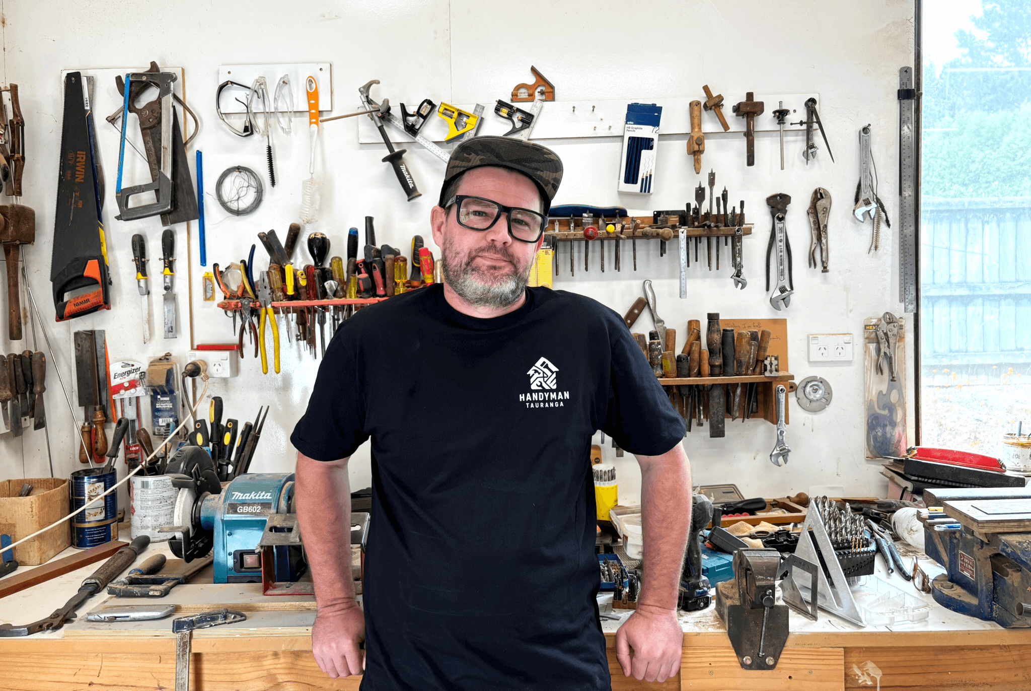 A man with glasses and a beard stands in a workshop surrounded by tools and equipment hanging on a white wall and placed on a workbench.