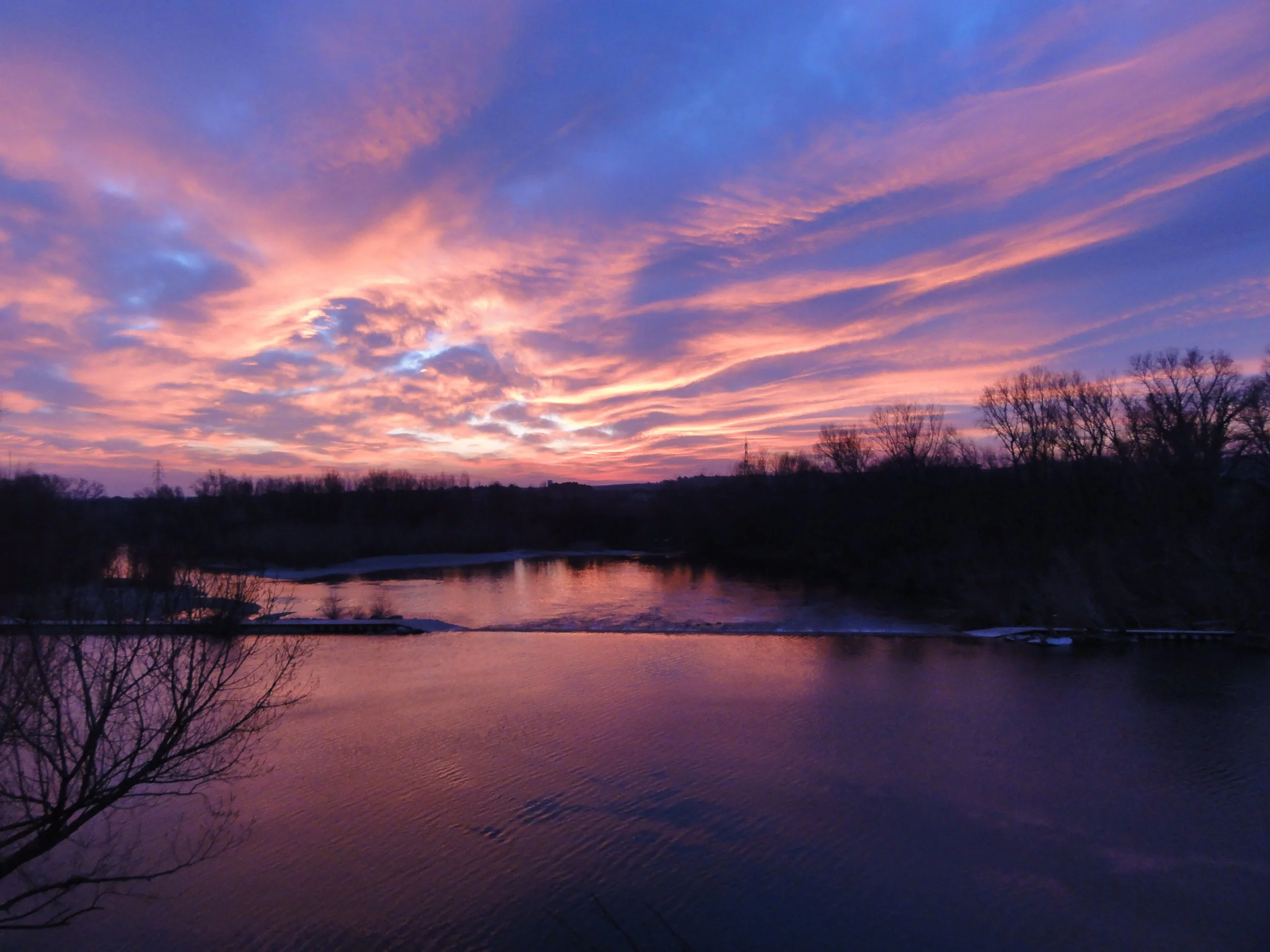 A colorful sunset over a river with trees silhouetted along the shoreline and pink and purple clouds in the sky.