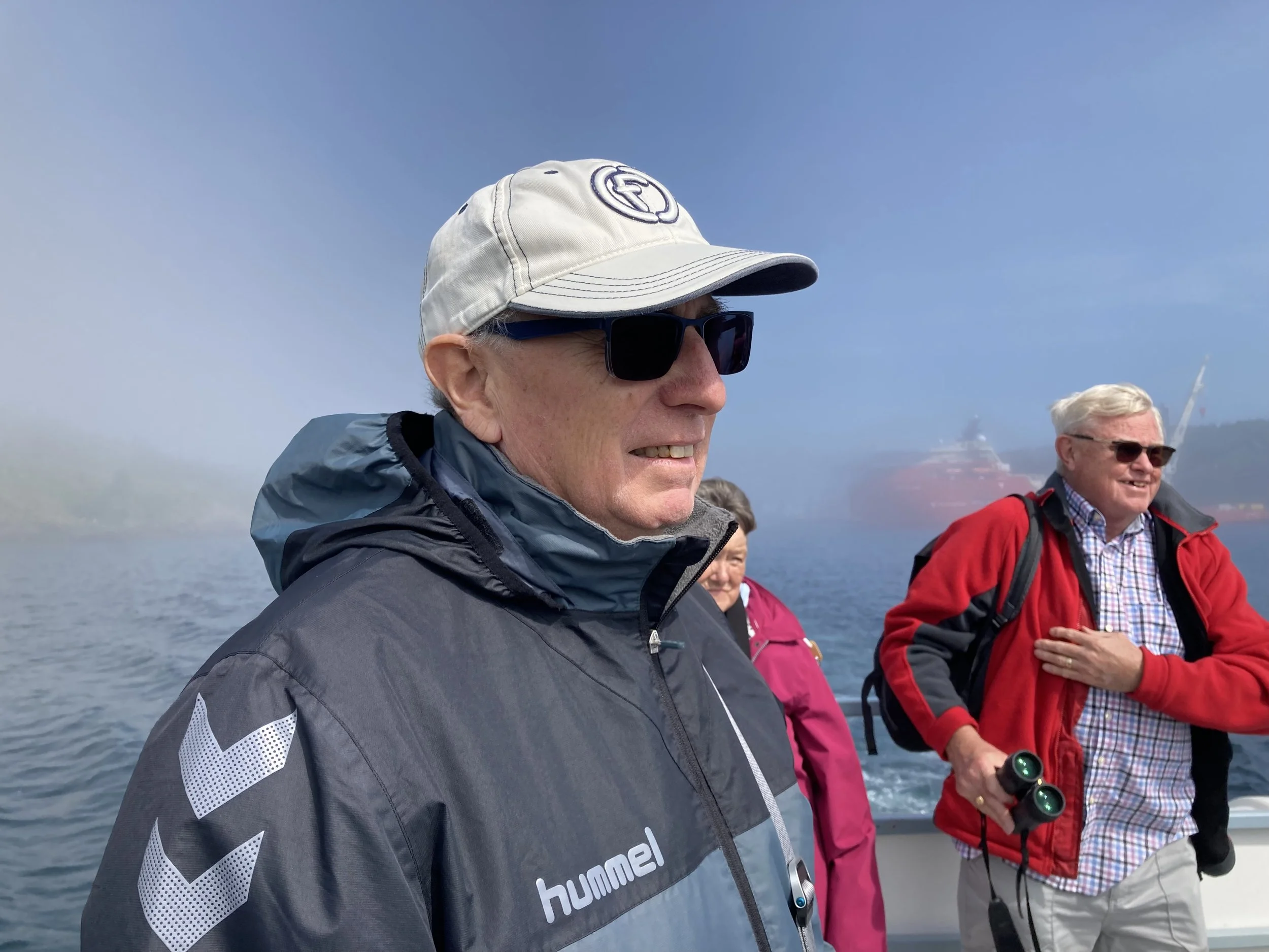 Group of people on a boat, with one man in the foreground wearing a gray jacket, white cap, and sunglasses, and two people behind him, one holding binoculars, all smiling. The scene is outdoors on the water with a foggy or cloudy background.