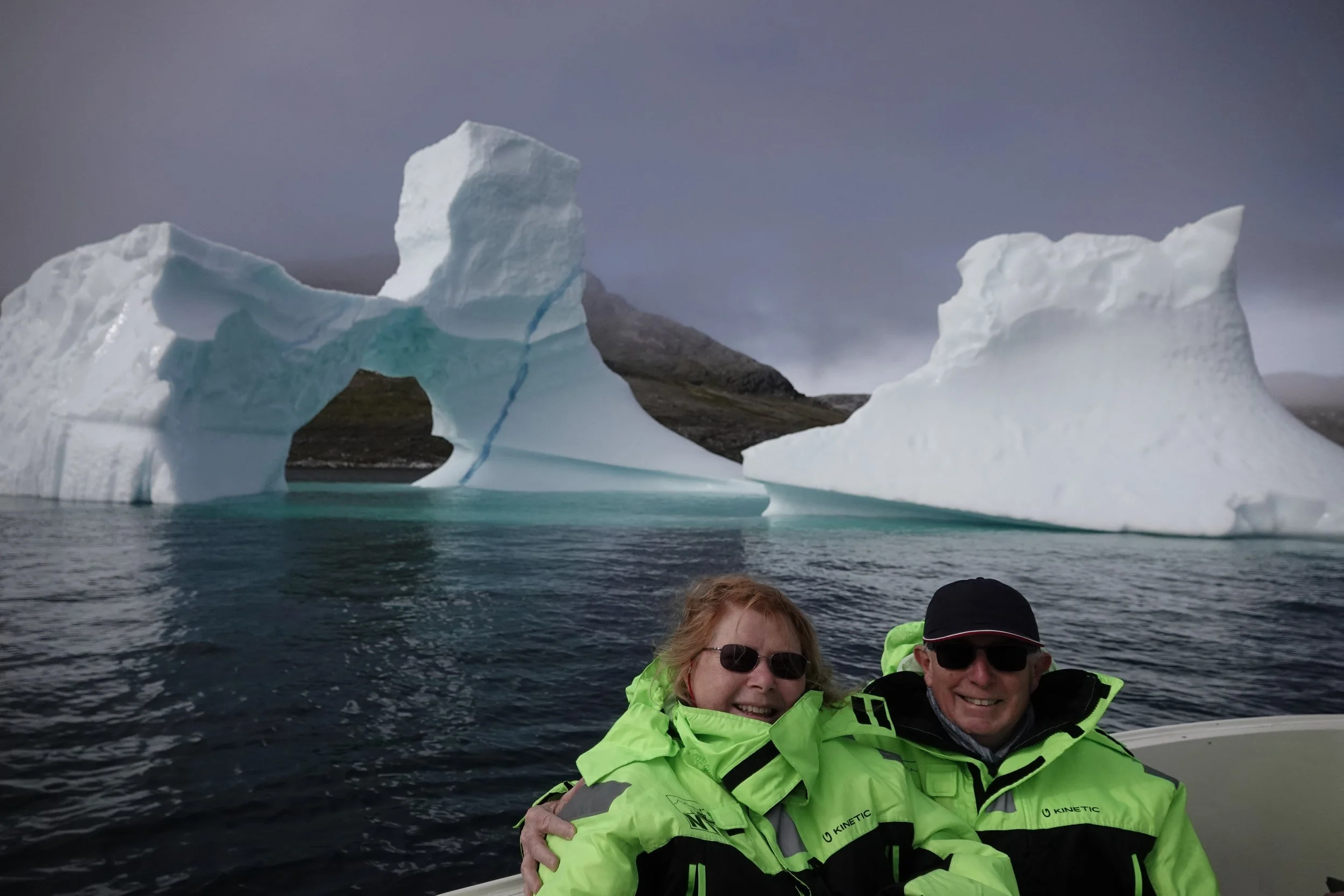 Two people in bright green jackets and sunglasses smiling on a boat, with large icebergs and a mountain in the background.