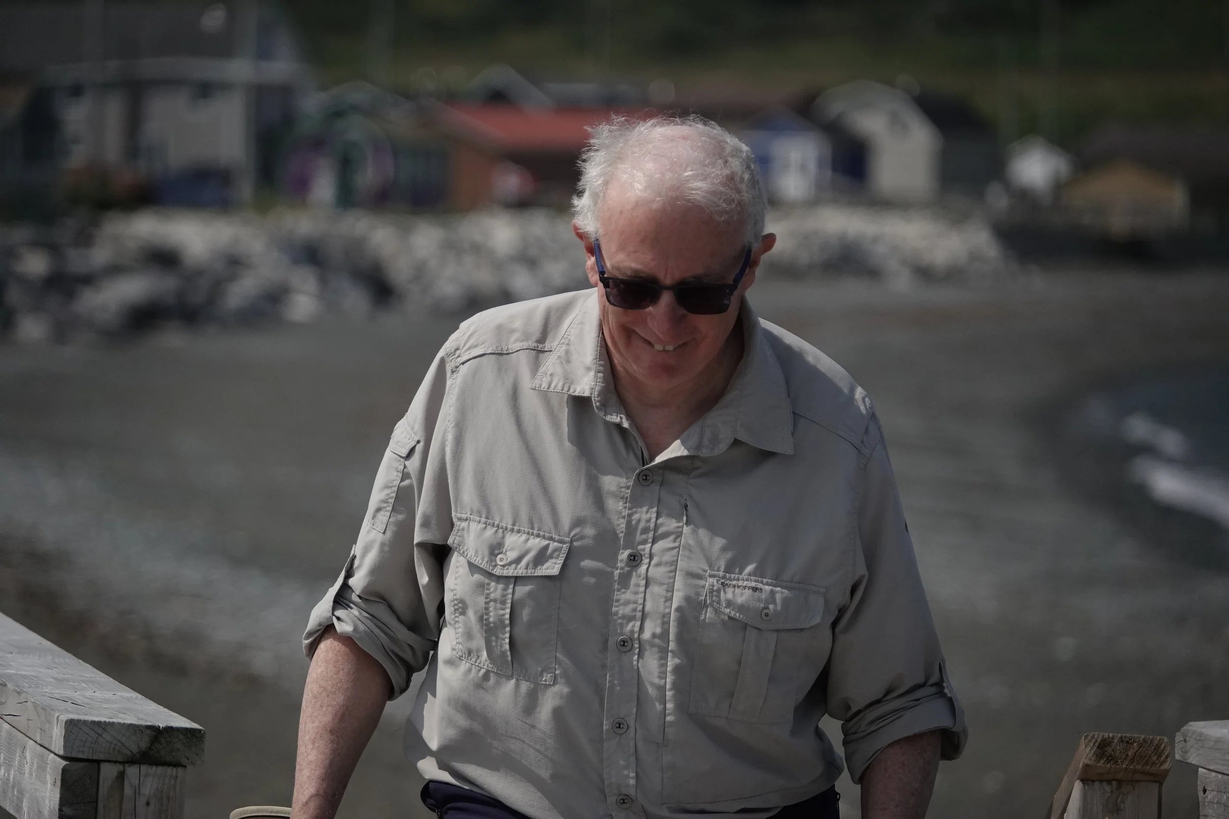 An older man with white hair and sunglasses, wearing a light beige shirt, smiling while walking on a beach with a wooden railing, with houses and rocky shoreline in the background.
