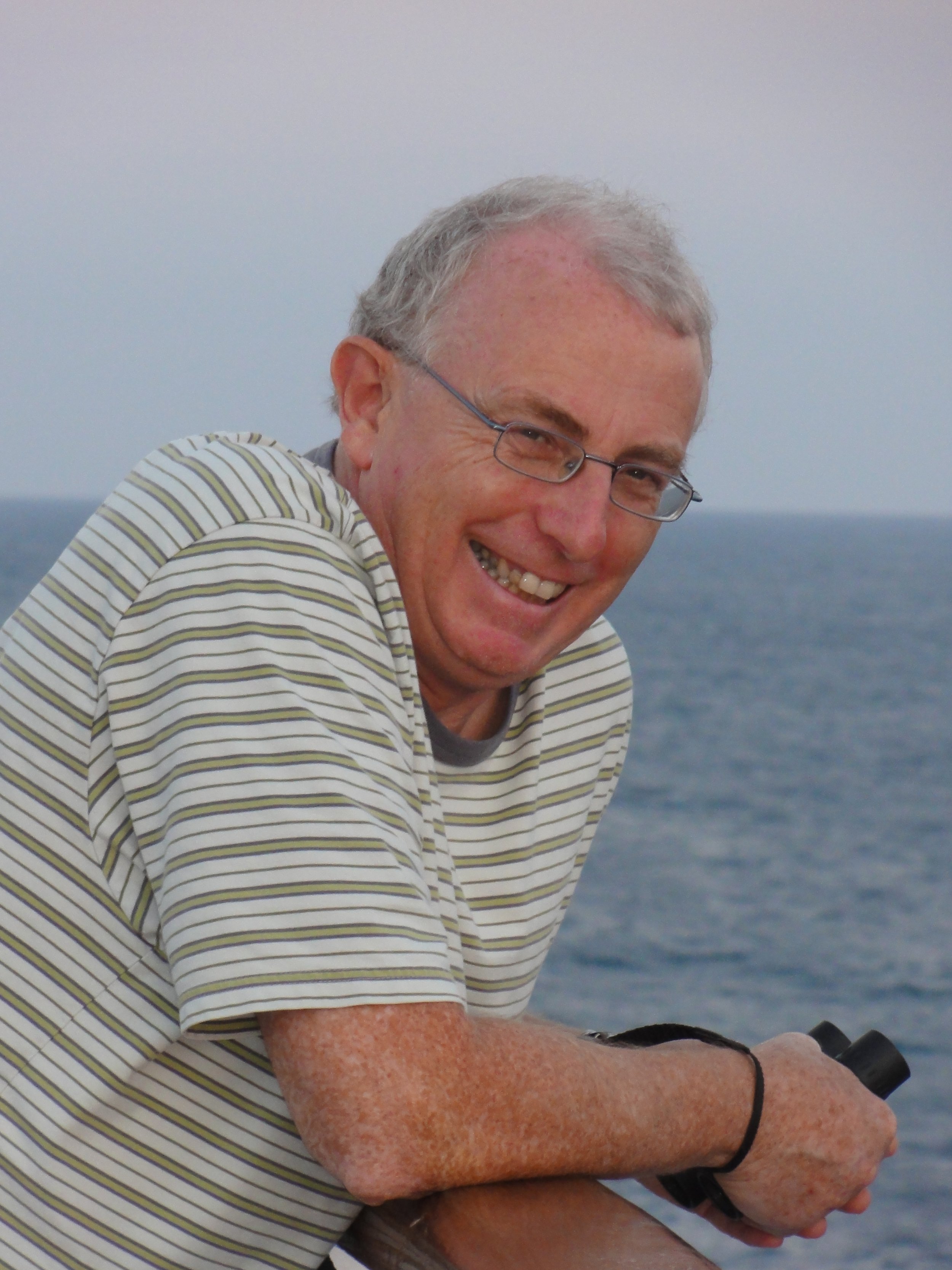 A smiling man with glasses and gray hair, wearing a striped shirt, looks at the camera while holding binoculars, with the ocean in the background.