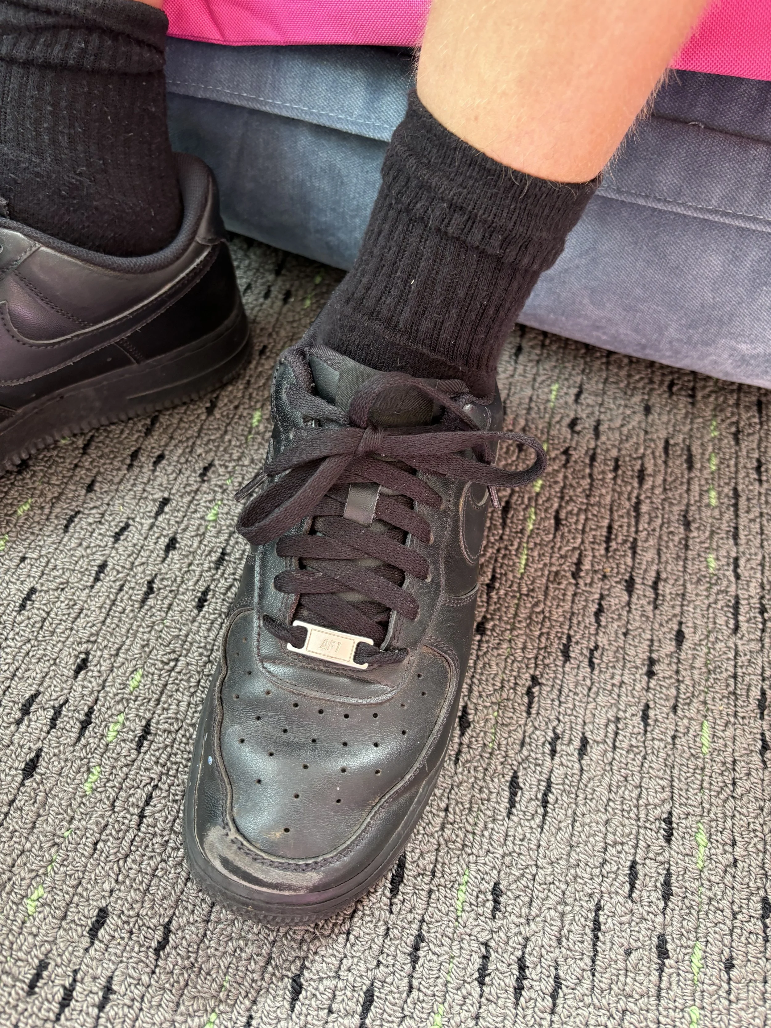 A black sneaker with black laces, worn with black socks, on a patterned carpeted floor next to another black shoe.