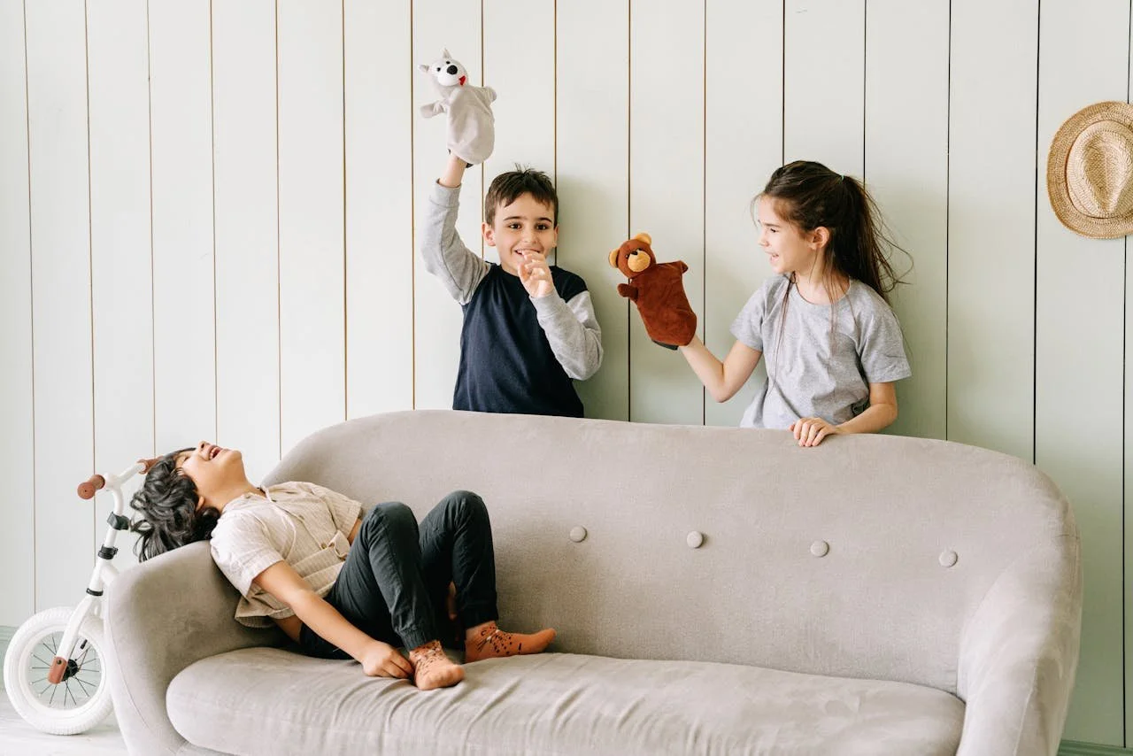 Two children are playing with sock puppets behind a sofa while a third child is lying on the sofa, laughing.
