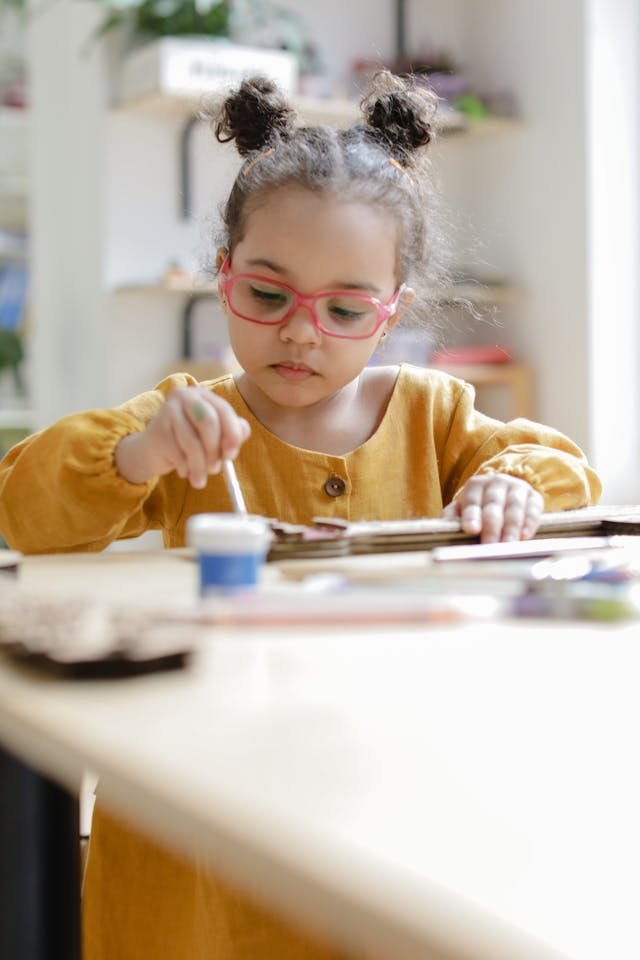 Young girl with red glasses painting or crafting at a table in a bright, organized room.