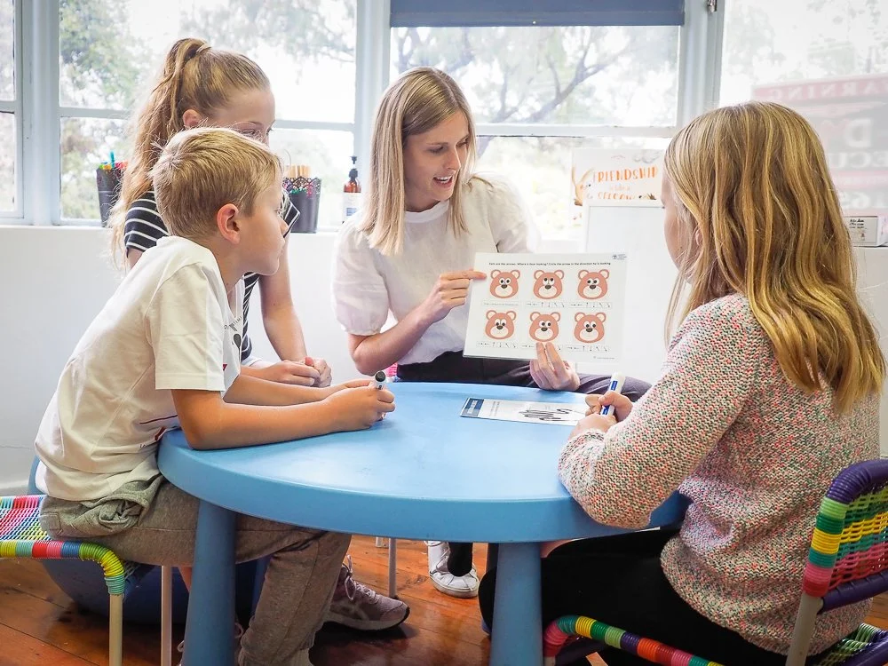 A teacher or therapist sitting at a round blue table with three children, using a picture chart with bear faces for an educational activity in a classroom.