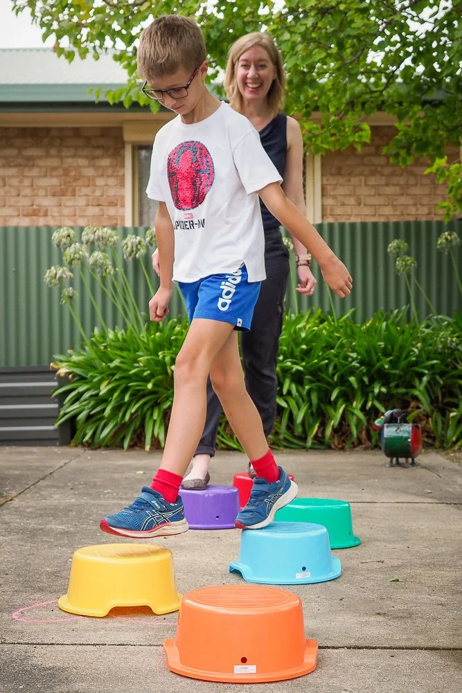 A young boy with glasses balancing on colorful upside-down buckets in a backyard, with a woman smiling nearby, surrounded by greenery and a brick house in the background.