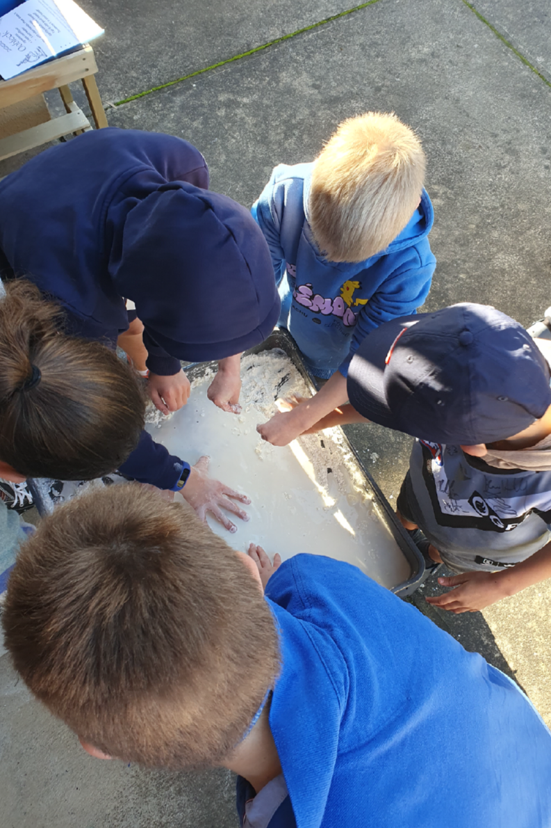 Children gathered around a tray of flour, mixing or playing with it outdoors.
