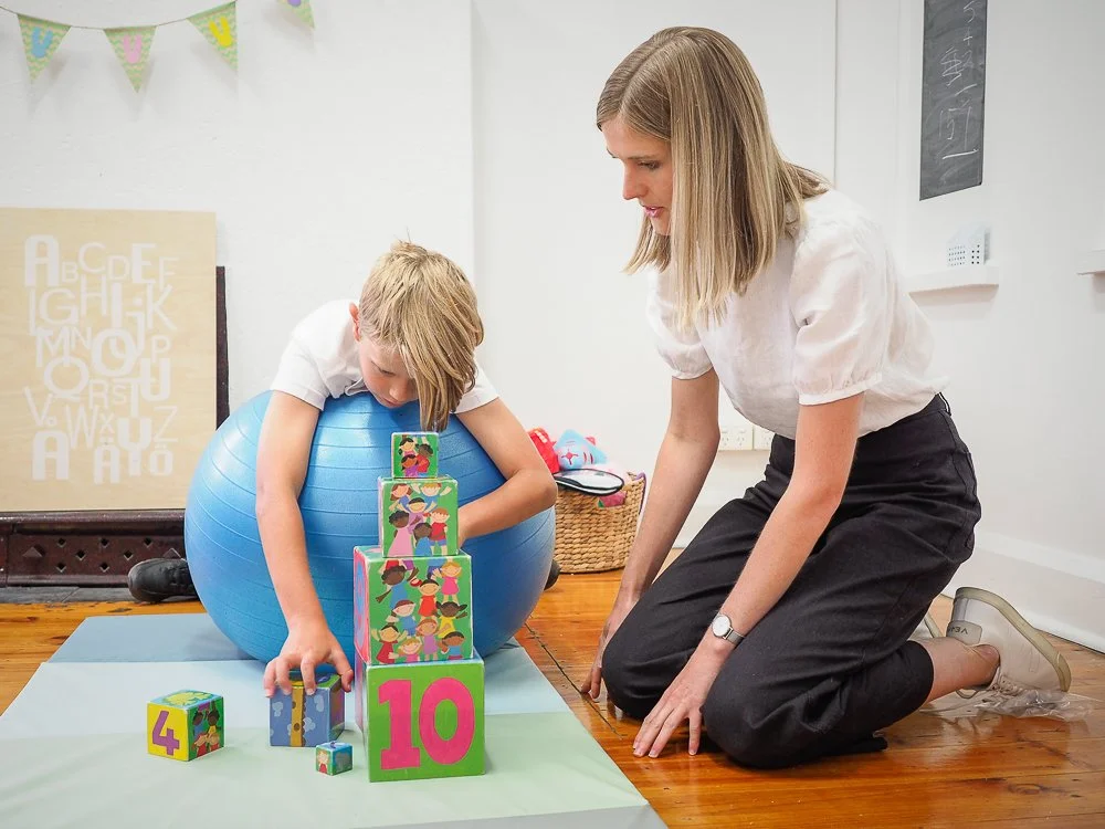 A woman and a young boy playing with colorful foam blocks and a blue exercise ball inside a room with white walls and wooden floor. The boy is leaning on the exercise ball while stacking blocks, and the woman is kneeling nearby.