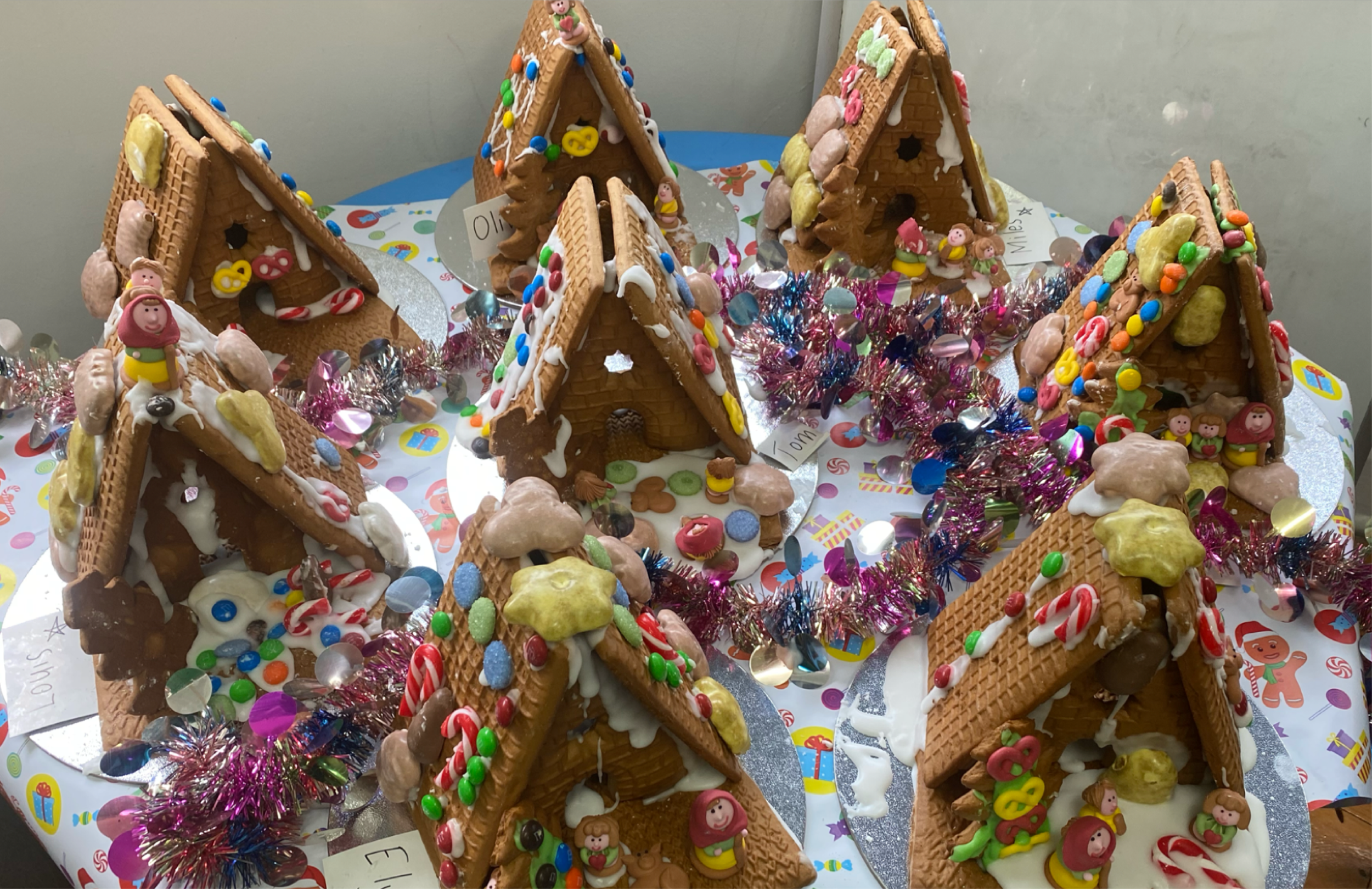 Gingerbread houses decorated with colorful candies, gumdrops, and icing, arranged around a festive table with shiny tinsel and holiday-themed wrapping paper.