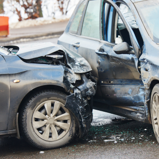 A rear-end collision between two cars with significant damage, including a crumpled front of a dark-colored vehicle and a damaged side of a light-colored SUV on a snowy road.