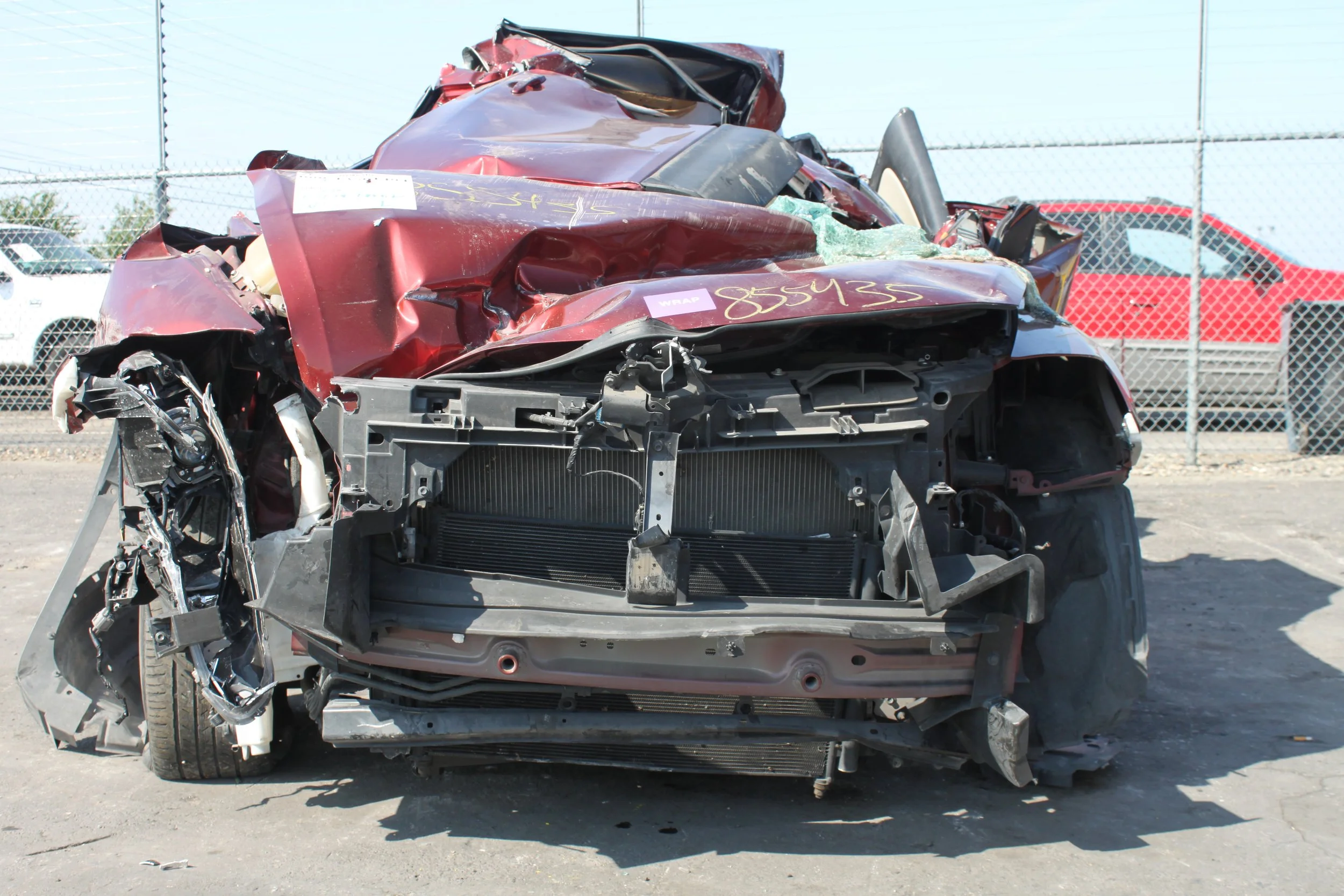 Damaged red car with front-end destruction in a salvage yard, surrounded by a chain-link fence.