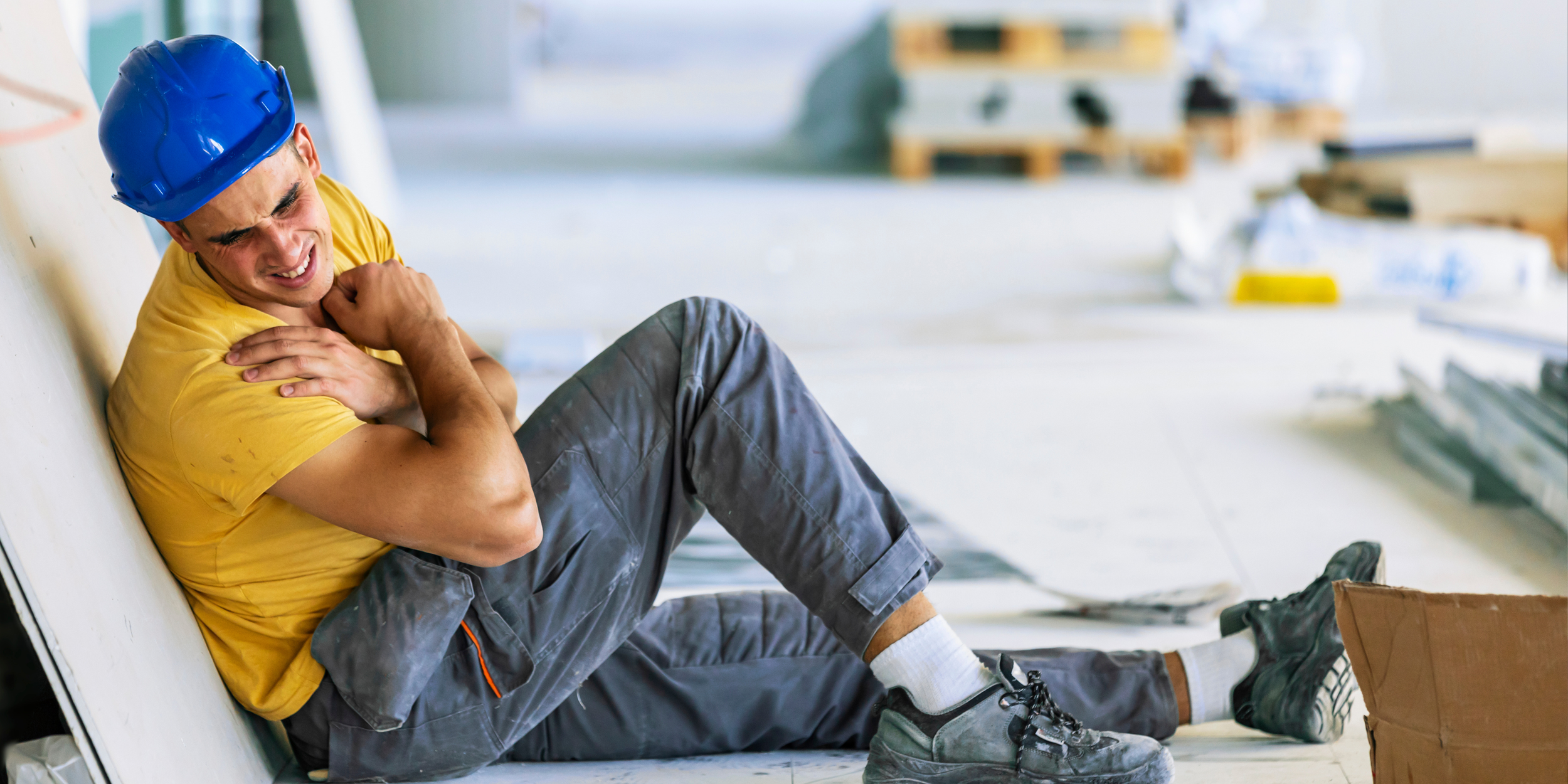 Construction worker wearing a blue hard hat and yellow t-shirt appears to be in pain, sitting on the floor with a hand on his neck at an indoor construction site.