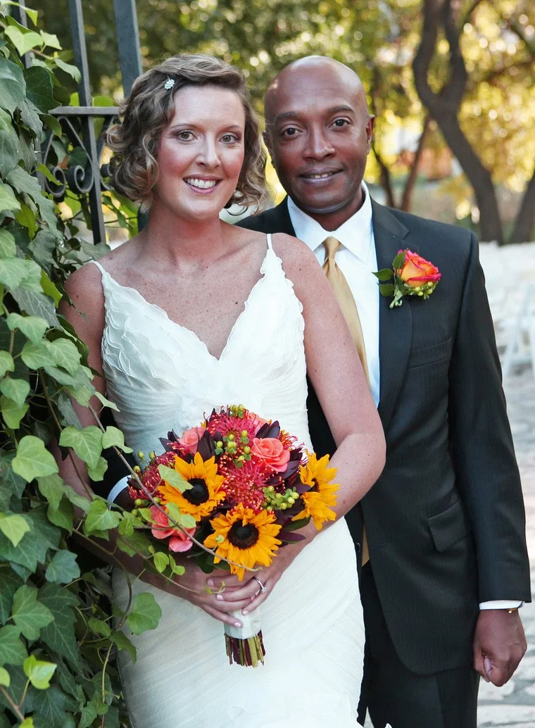 A newlywed couple standing outdoors, the bride holding a colorful bouquet of sunflowers, roses, and greenery, while the groom wears a black suit with a white shirt, yellow tie, and a boutonniere matching the bride's bouquet. They are surrounded by greenery and trees.