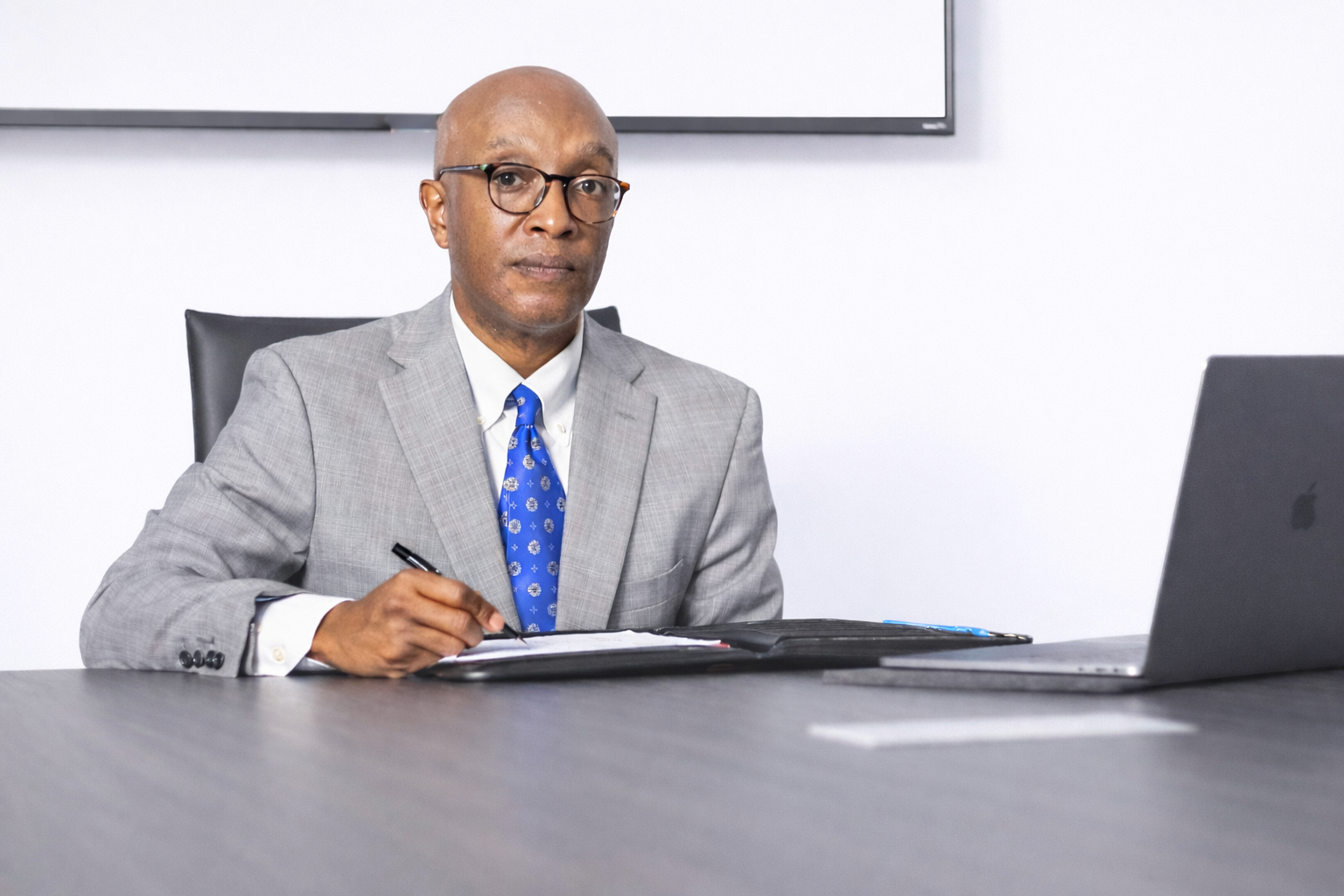 A professional black San Antonio lawyer in a gray suit and blue tie sitting at a conference table with a laptop and notepad, looking at the camera.