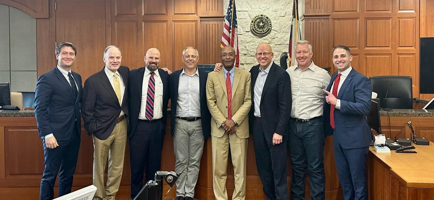 Group of nine men standing together in a courtroom or government room, dressed in business attire, with wood-paneled walls, American flags, and official symbols in the background.