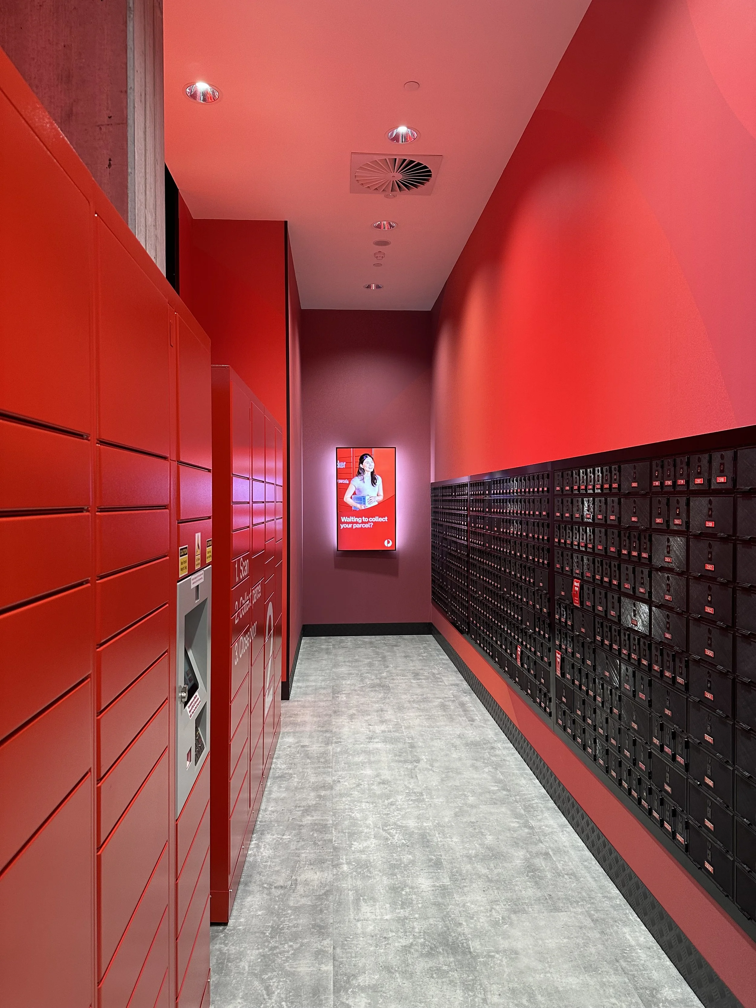 Red parcel lockers and black mailboxes in a modern corridor with a digital screen at the end.