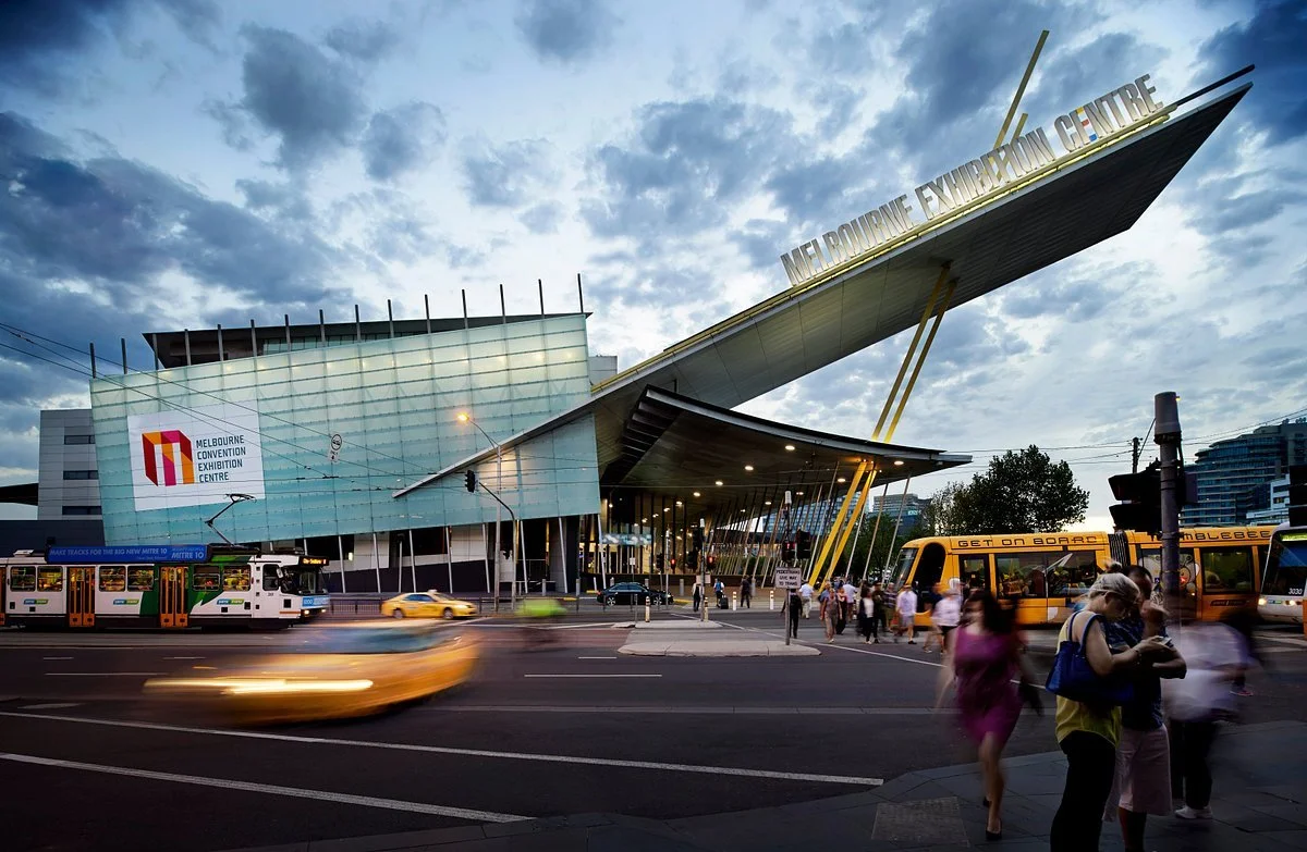 The Melbourne Convention and Exhibition Centre with a unique angular modern architectural design, moving vehicles, and people crossing the street during dusk.