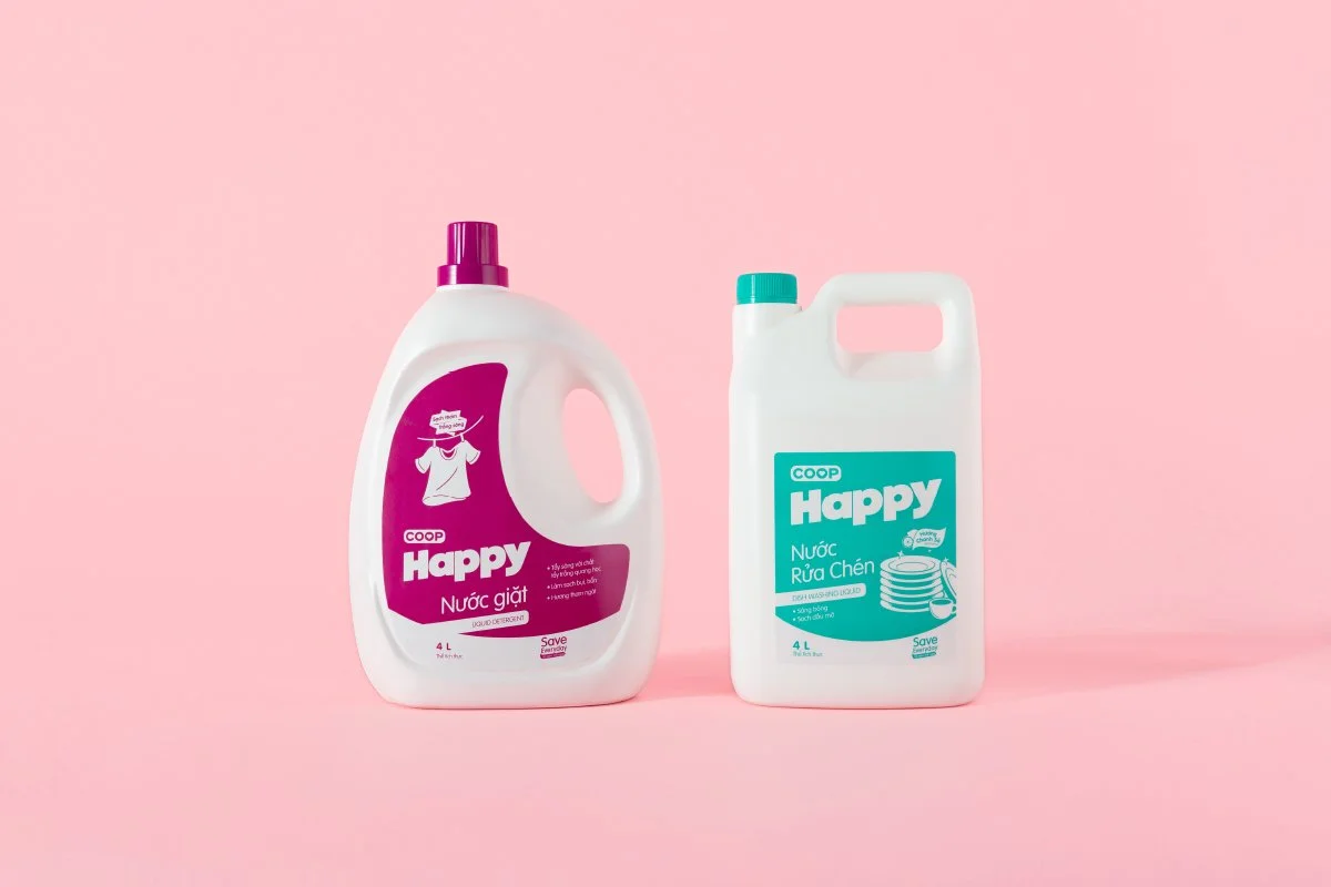 Two bottles of cleaning liquids on a pink background, labeled 'COOP Happy'. The left bottle is a liquid detergent, and the right bottle is dishwashing liquid.