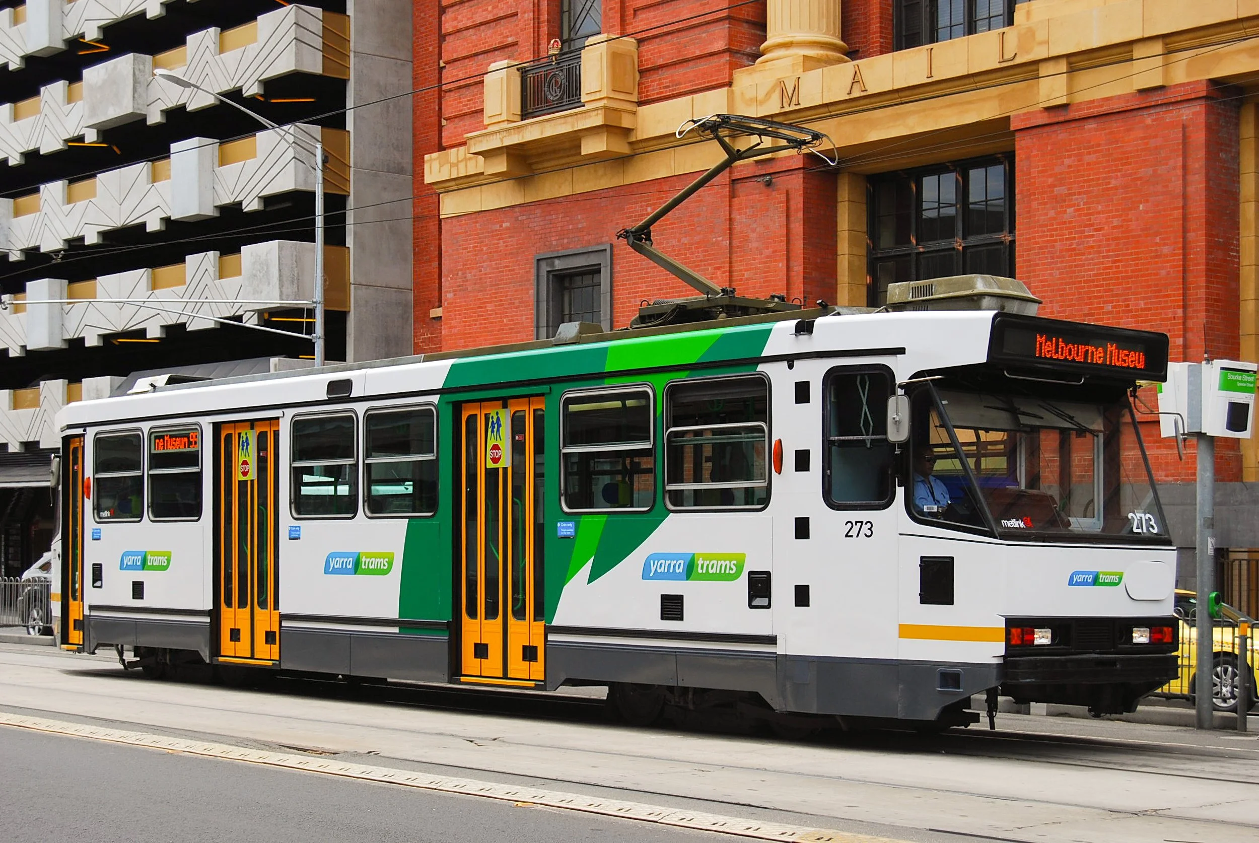 Modern tram on city street with brick and concrete buildings in background, displaying 'Melbourne Museum' on digital sign.