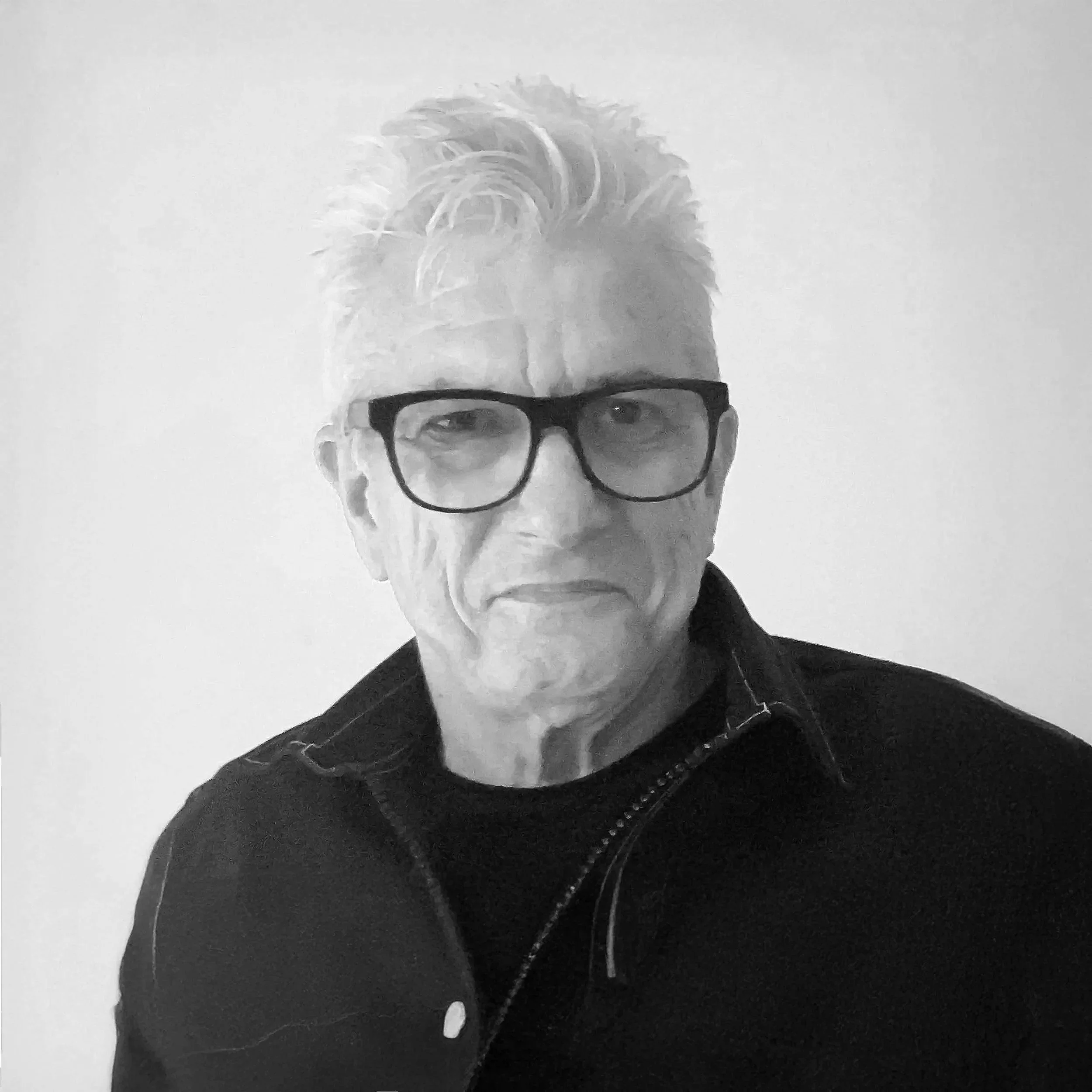 Black and white portrait of an older man with glasses, short wavy hair, wearing a dark zip-up sweater.