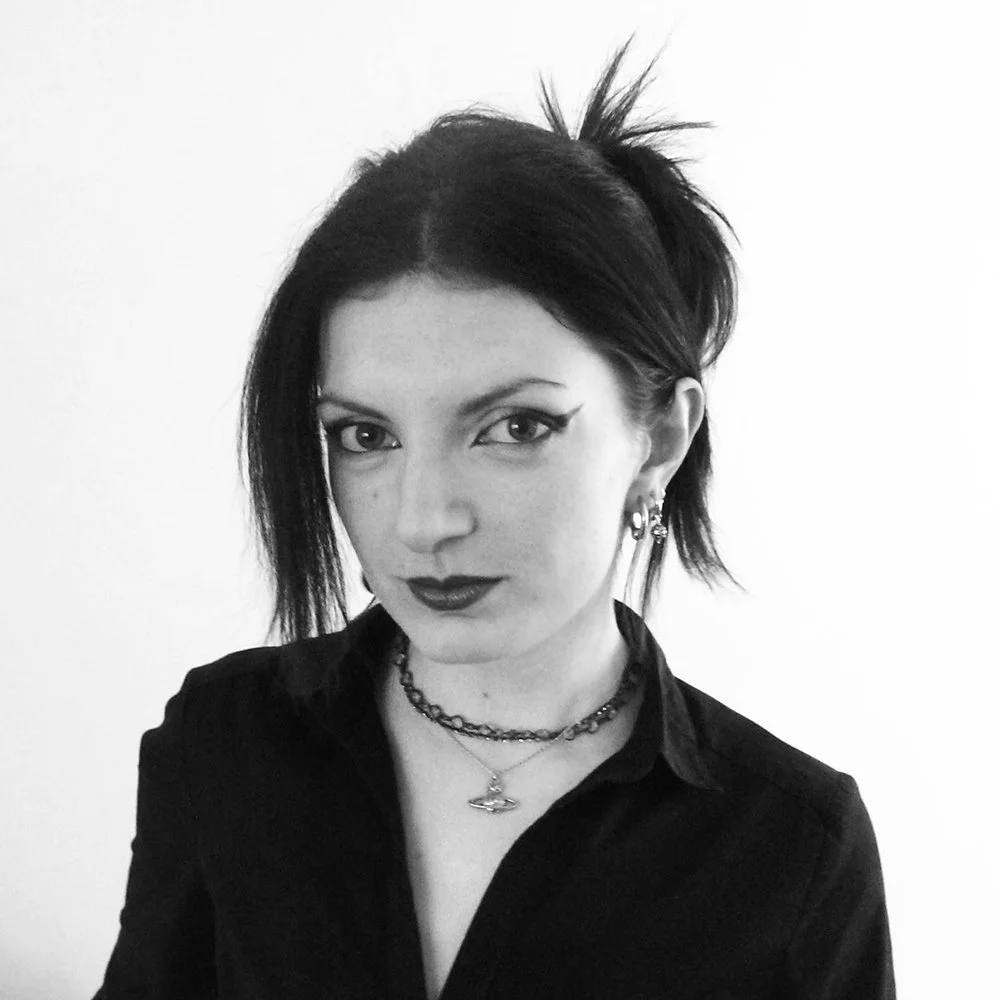Black and white photo of a woman with dark, spiky hair, wearing dark lipstick, eyeliner, layered necklaces, and earrings, against a plain light background.