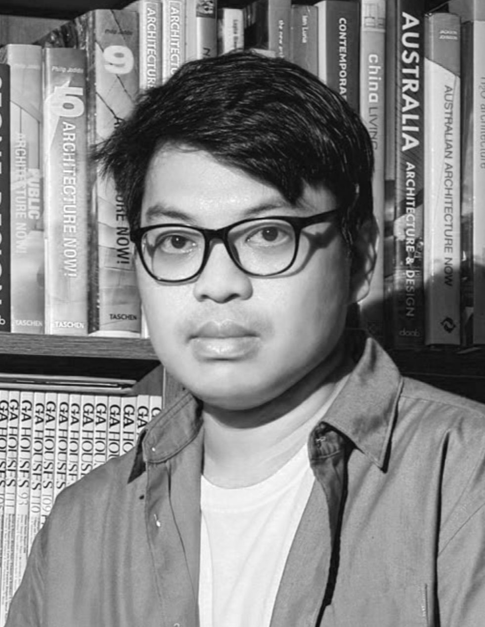 A black-and-white photo of a young man with dark hair and glasses, standing in front of a bookshelf filled with architecture books.