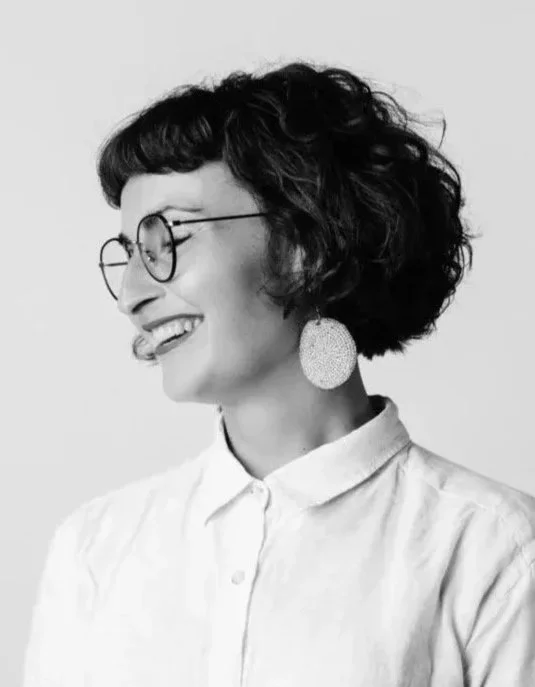 Black and white photo of a woman with short, curly hair, wearing glasses, round earrings, and a collared shirt, smiling and looking to the side.