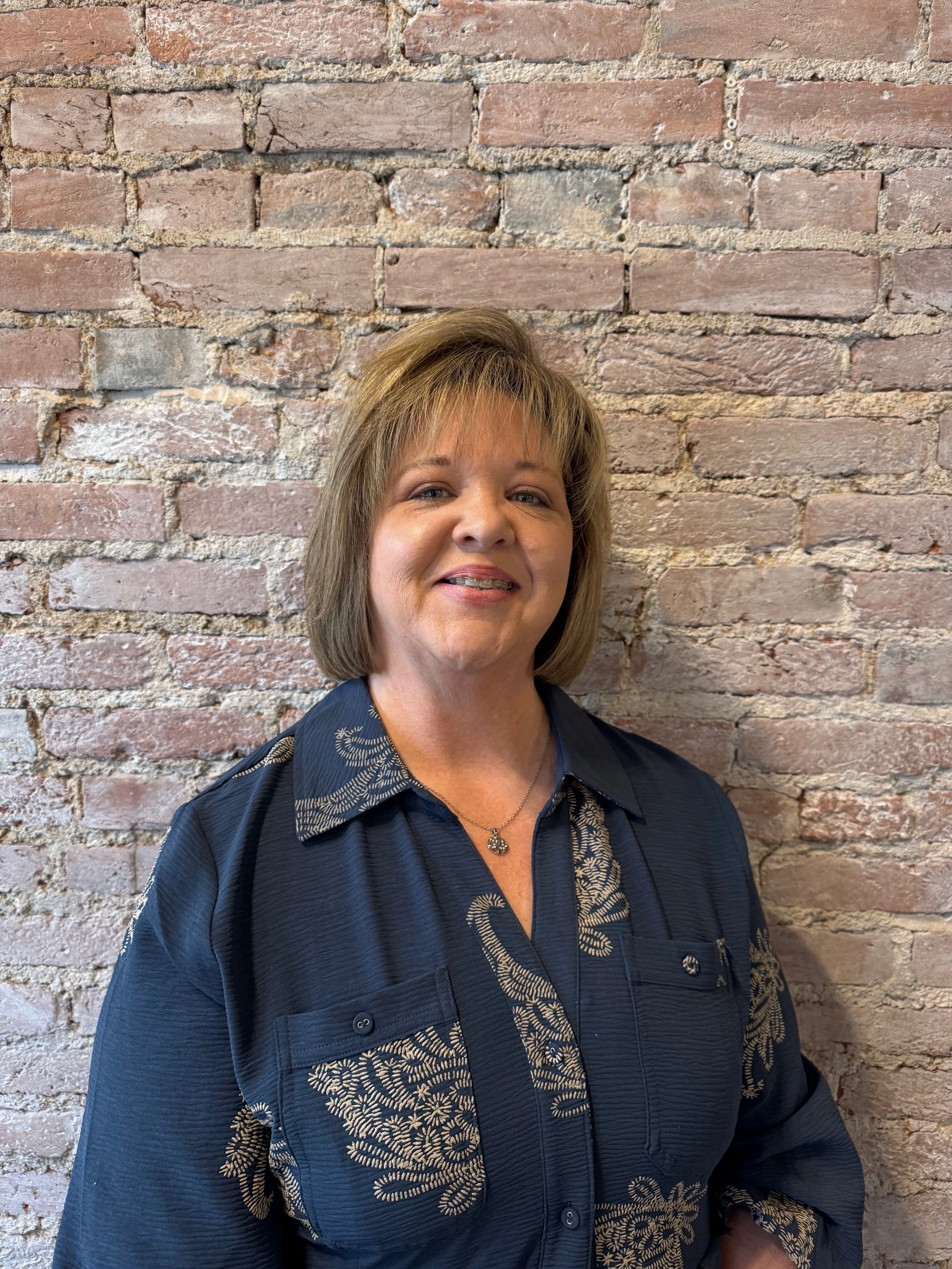 A woman with short, light brown hair smiling, wearing a dark blue shirt with beige embroidery, standing in front of an exposed brick wall.