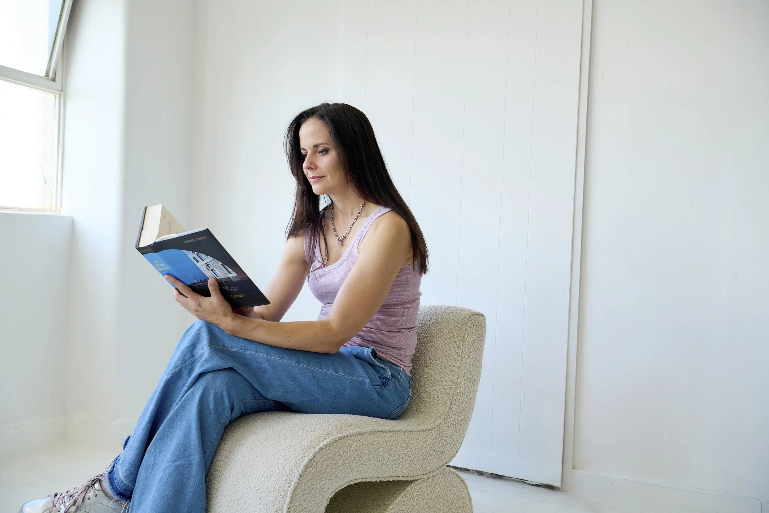 Woman sitting on a curved beige chair reading a book in a bright room with white walls and a window.