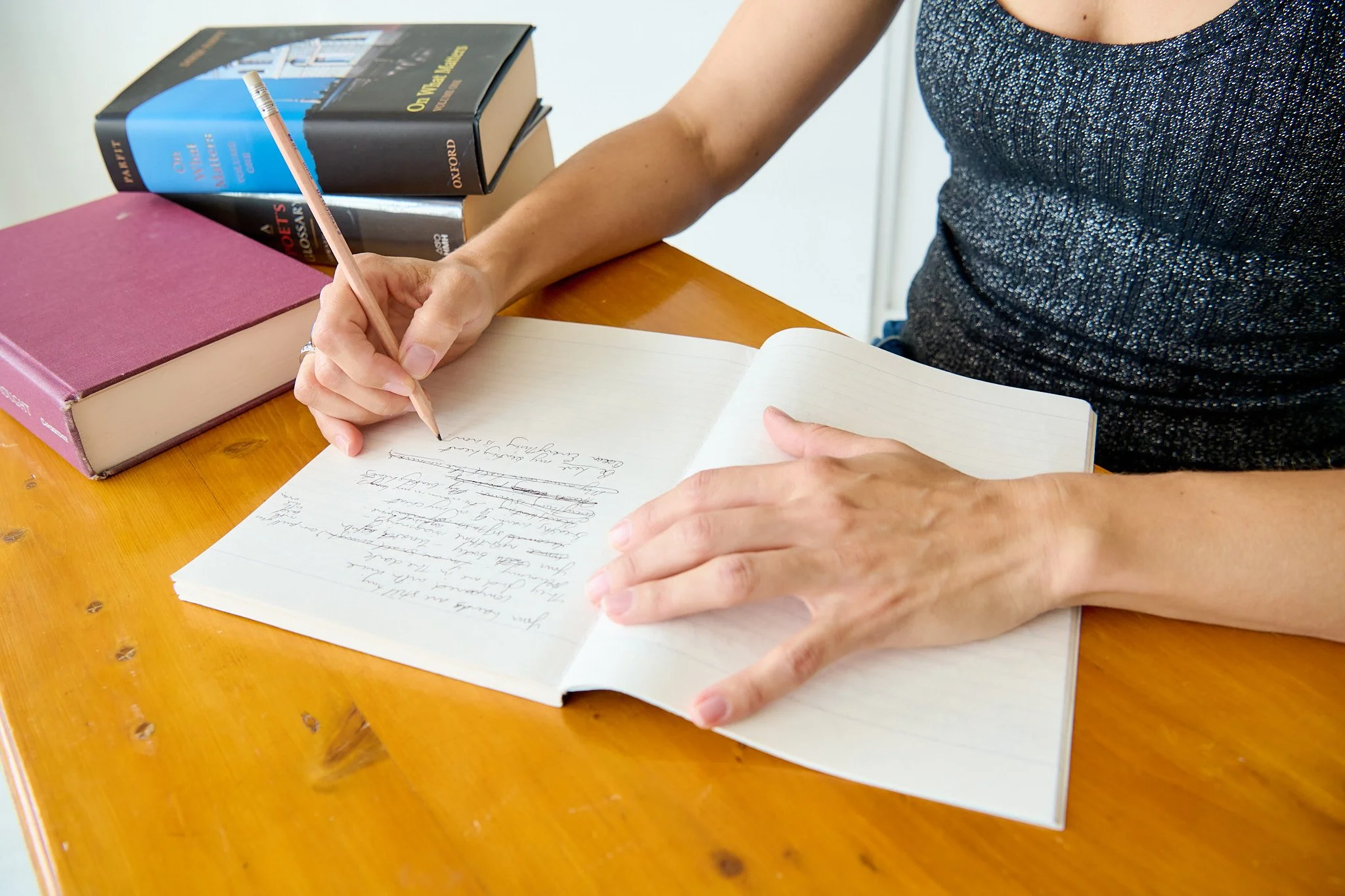 A person is writing in a notebook with a pink pencil at a wooden table. There are stacked books nearby, including a large pink book and several larger, blue, black, and gray books.