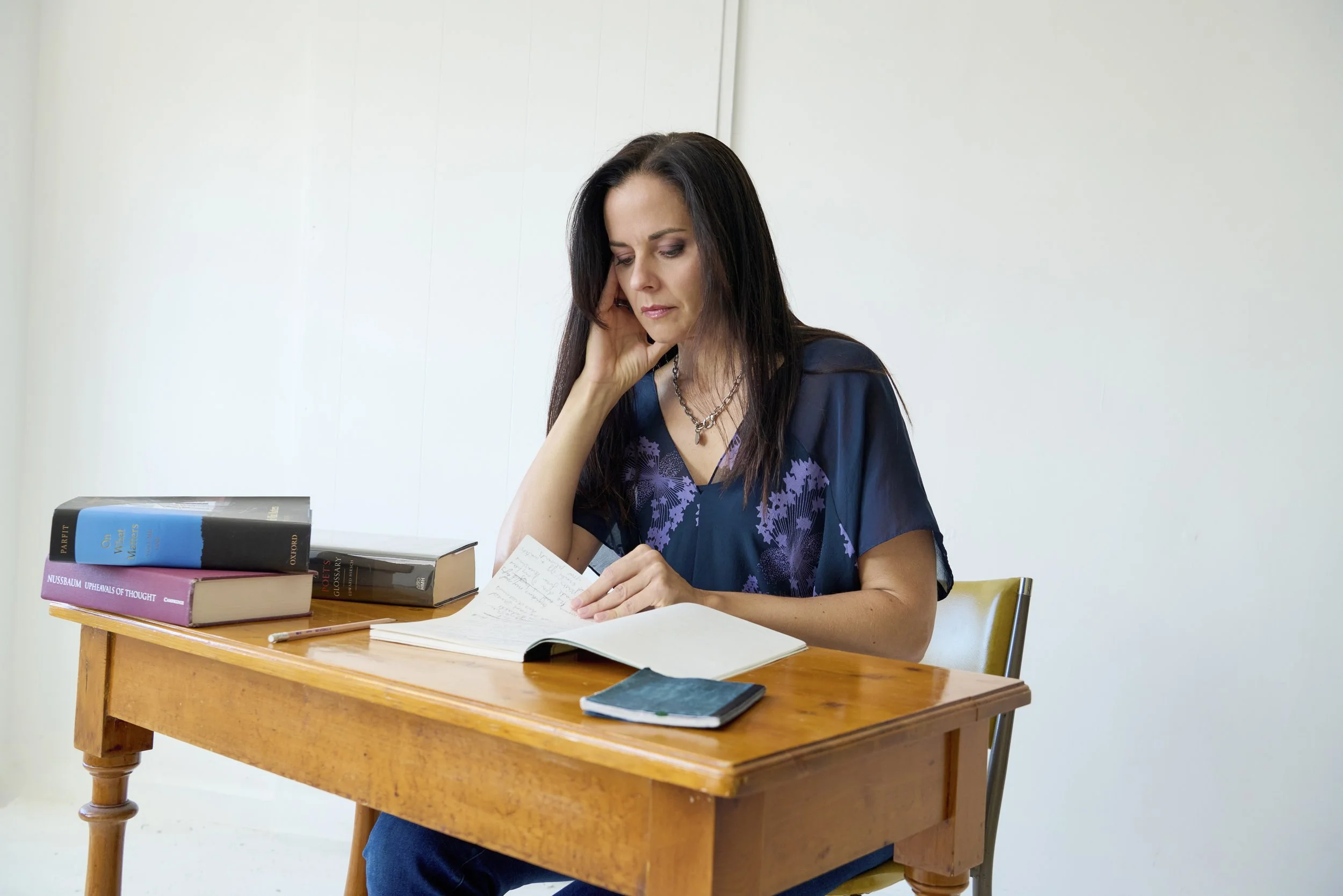 A woman with long dark hair sitting at a wooden table, reading and writing in a notebook, with several books and a phone in front of her, in a plain white room.