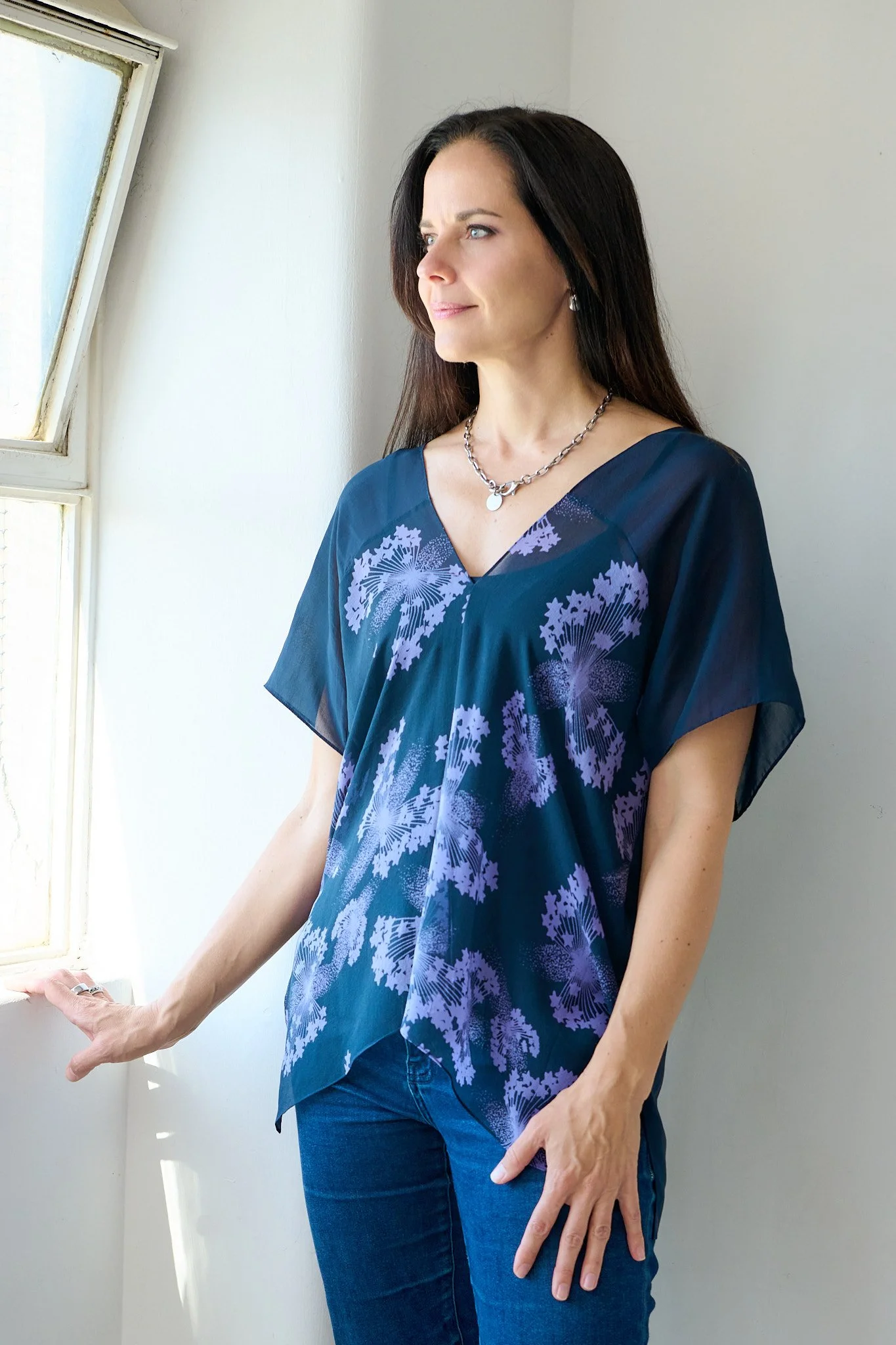 A woman with long dark hair wearing a blue patterned top and jeans, standing indoors near a window, looking thoughtfully outside.