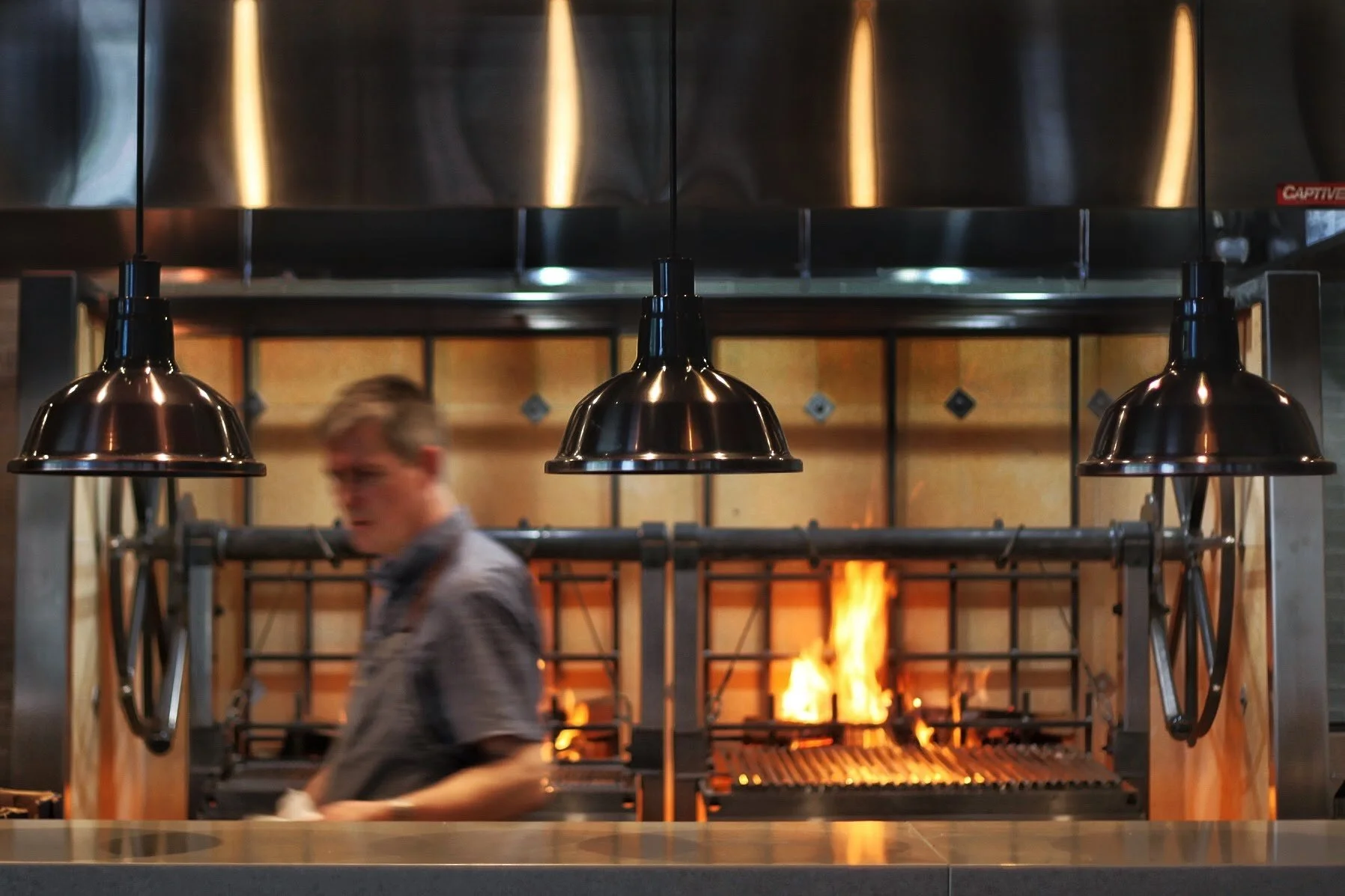 A blurry man working in a restaurant kitchen with a grill and open flame in the background, viewed through three hanging kitchen lights.