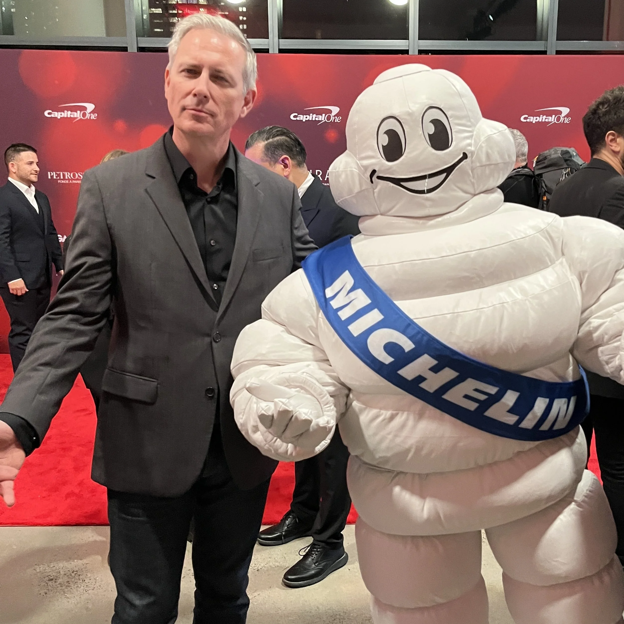 Chef Andrew Zimmerman in a dark suit posing next to the Michelin Man mascot with a blue sash that says 'MICHELIN' at an event with a red carpet background and other people in the background.