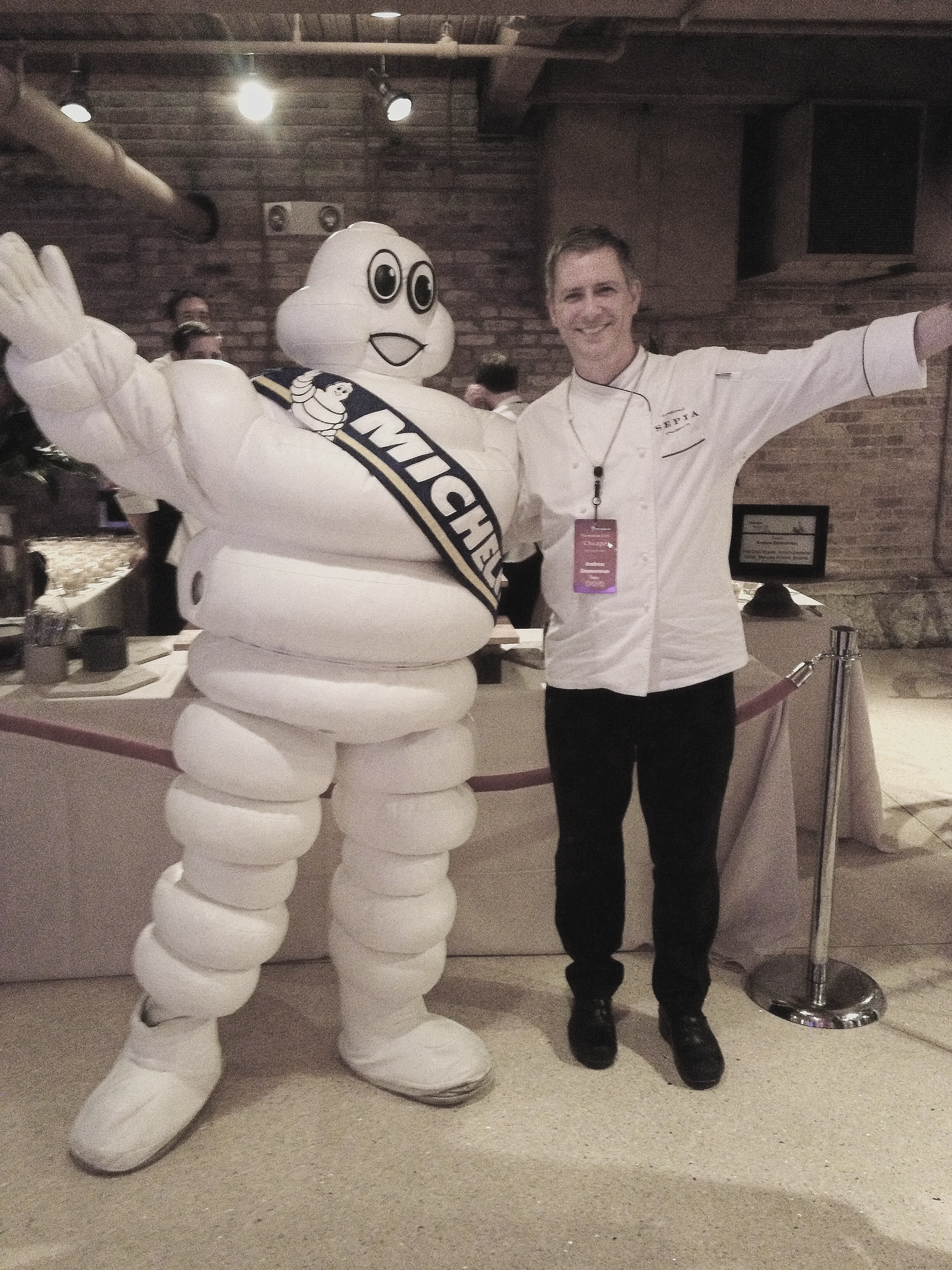 Chef Andrw Zimmerman dressed in a chef's uniform posing with the Michelin Man mascot at an event.