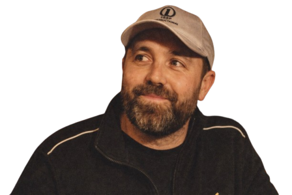 A man with a beard wearing a beige cap and black zip-up jacket, looking to his left.